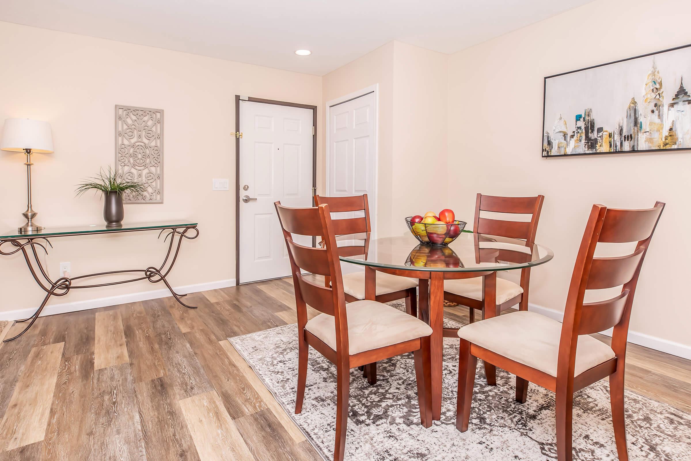 A dining area featuring a round glass table surrounded by four wooden chairs. On the table, there is a bowl of colorful fruits. The room has light-colored walls, a decorative wall piece, and a small console table with a plant. Natural light illuminates the space, creating a warm atmosphere.