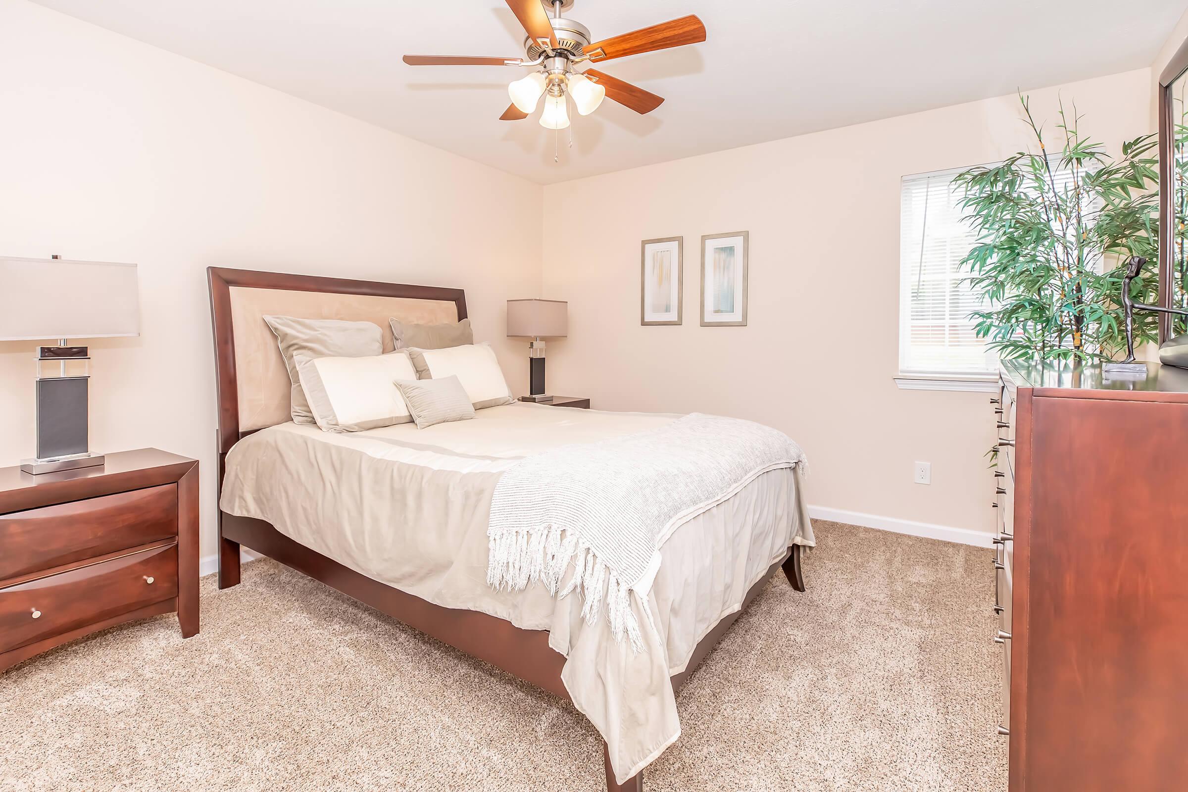 A cozy bedroom featuring a queen-sized bed with neutral bedding, a wooden headboard, and matching nightstands with lamps. A dresser is positioned against the wall, and a small window allows natural light. The room has soft beige walls and plush carpet, creating a warm and inviting atmosphere. A potted plant adds a touch of greenery.