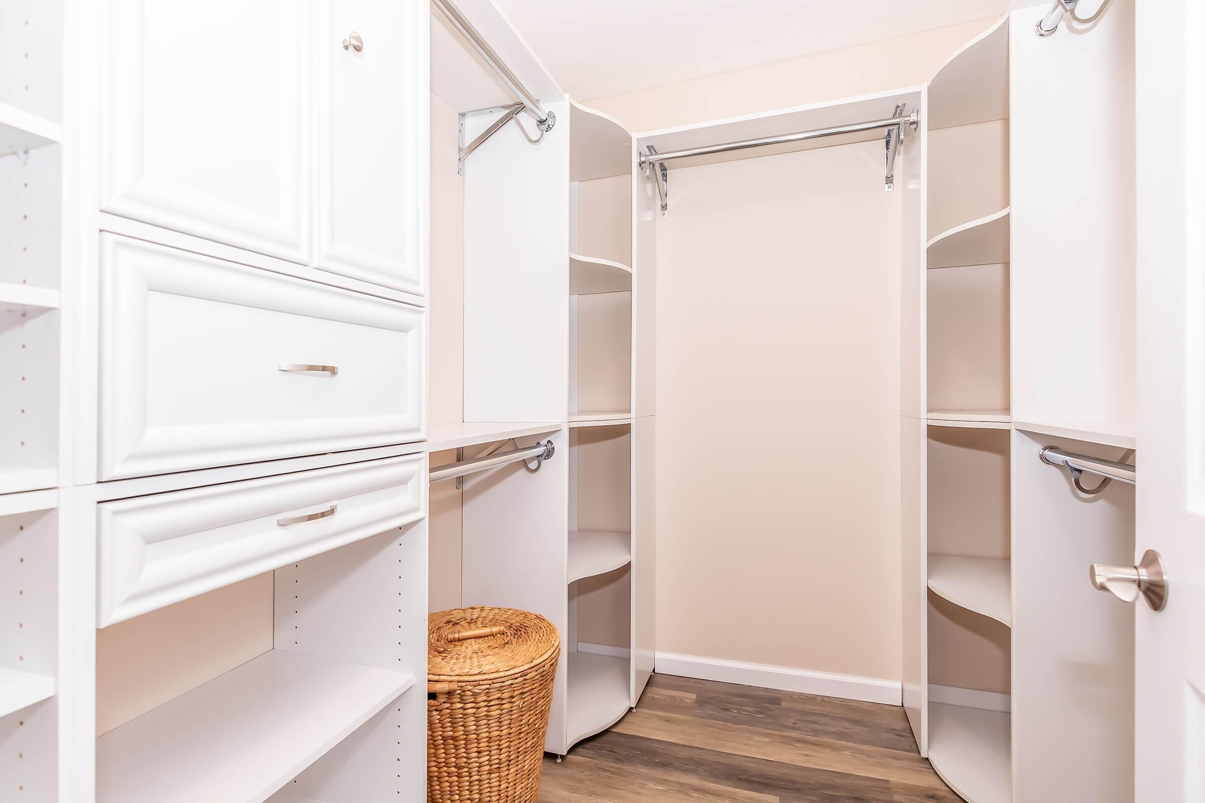 A spacious, organized closet interior featuring white shelving units, hanging rods, and a wicker basket on the floor. The walls are a light color, and the flooring is wooden, creating a clean and minimalist storage space.