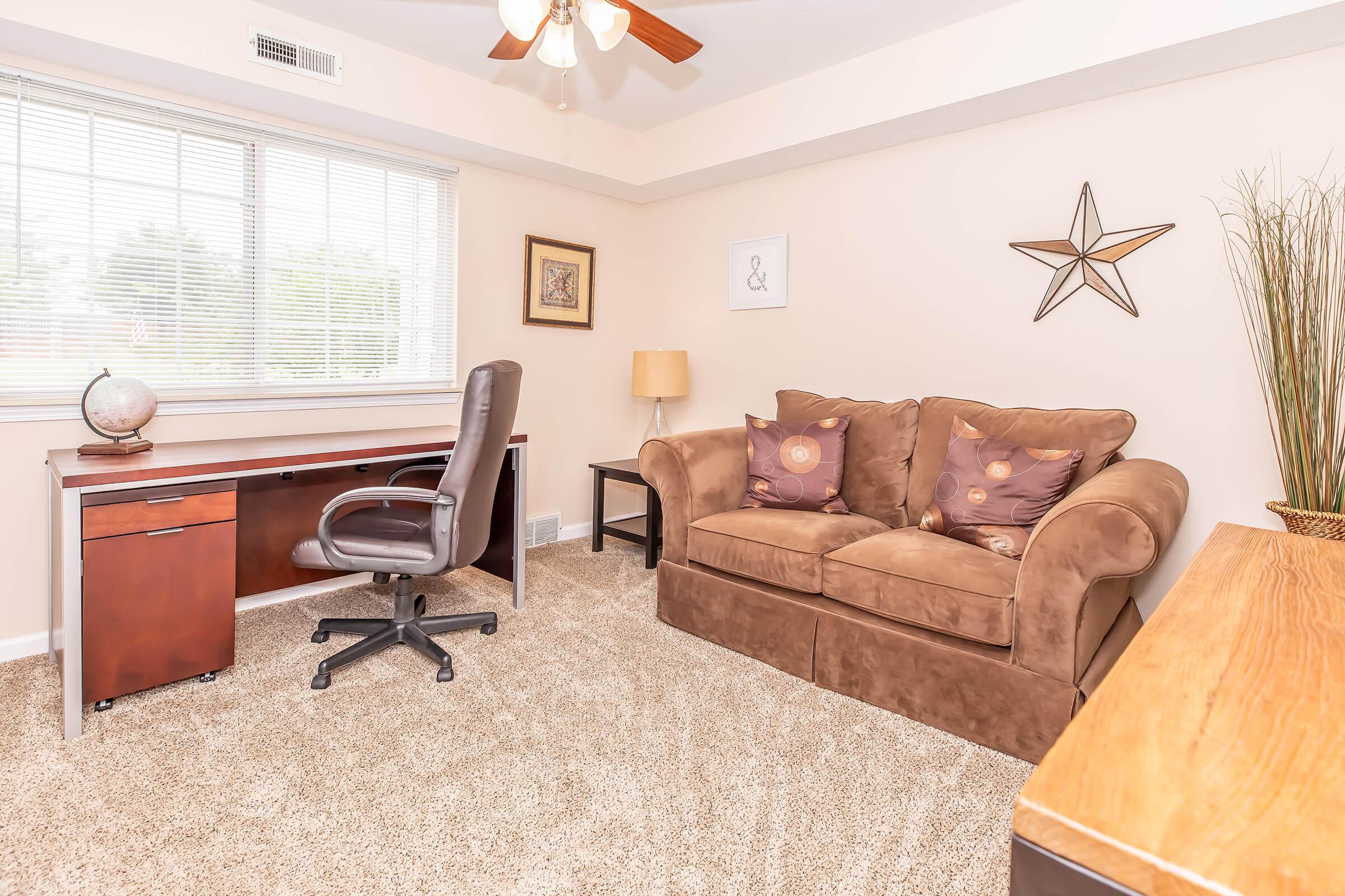 A cozy room featuring a brown loveseat with decorative cushions, a wooden desk with a rolling chair, a lamp, and a decorative star on the wall. The large window allows natural light to fill the space, with a globe accent on the desk and a potted plant in the corner, creating a welcoming atmosphere.