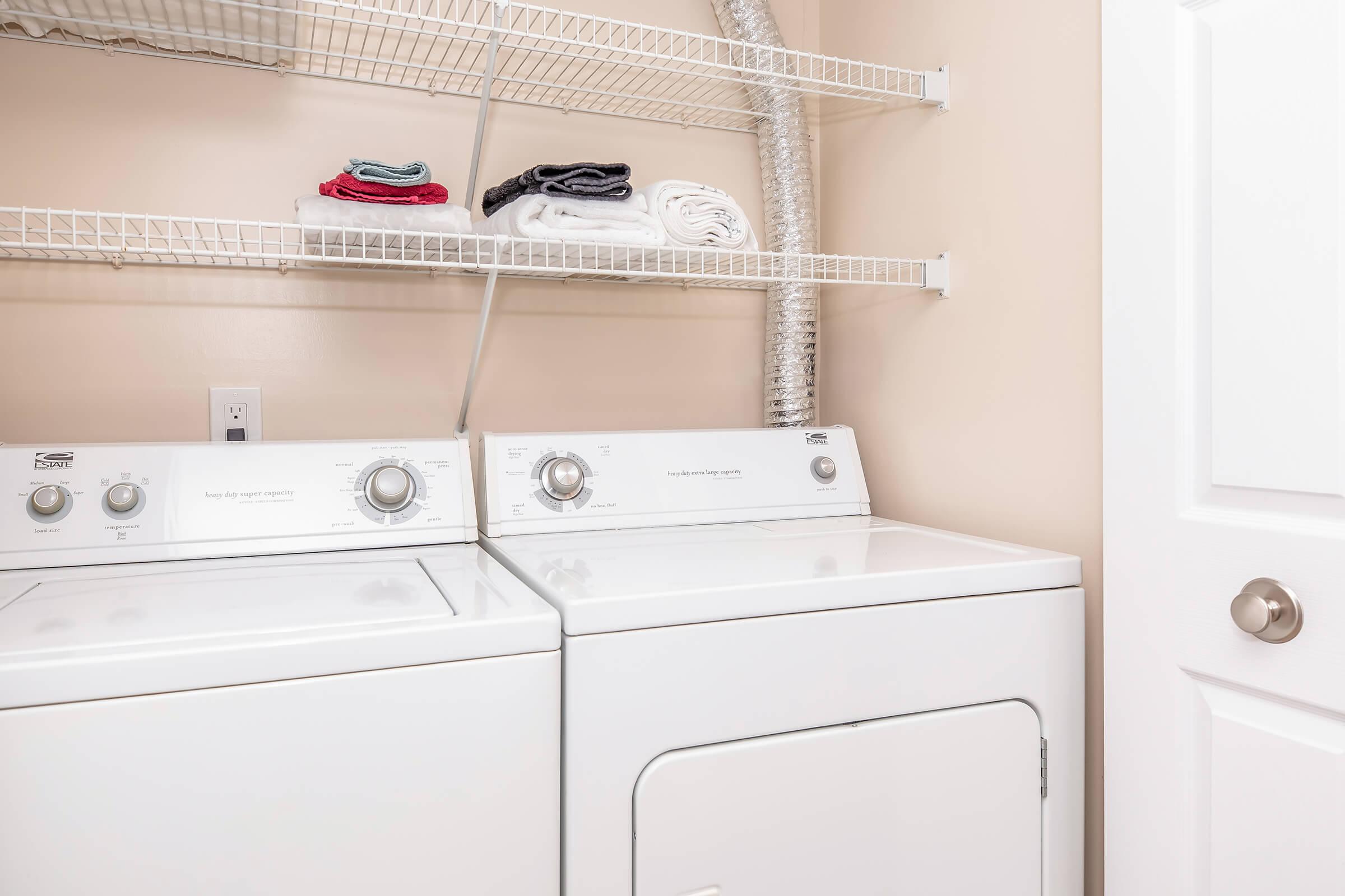 A laundry room featuring a stacked washer and dryer in white, with a metal vent hose attached. Above the appliances, there are neatly folded towels and cloths on a wire shelf. The walls are painted a light beige, and a door is visible on the right side of the image.