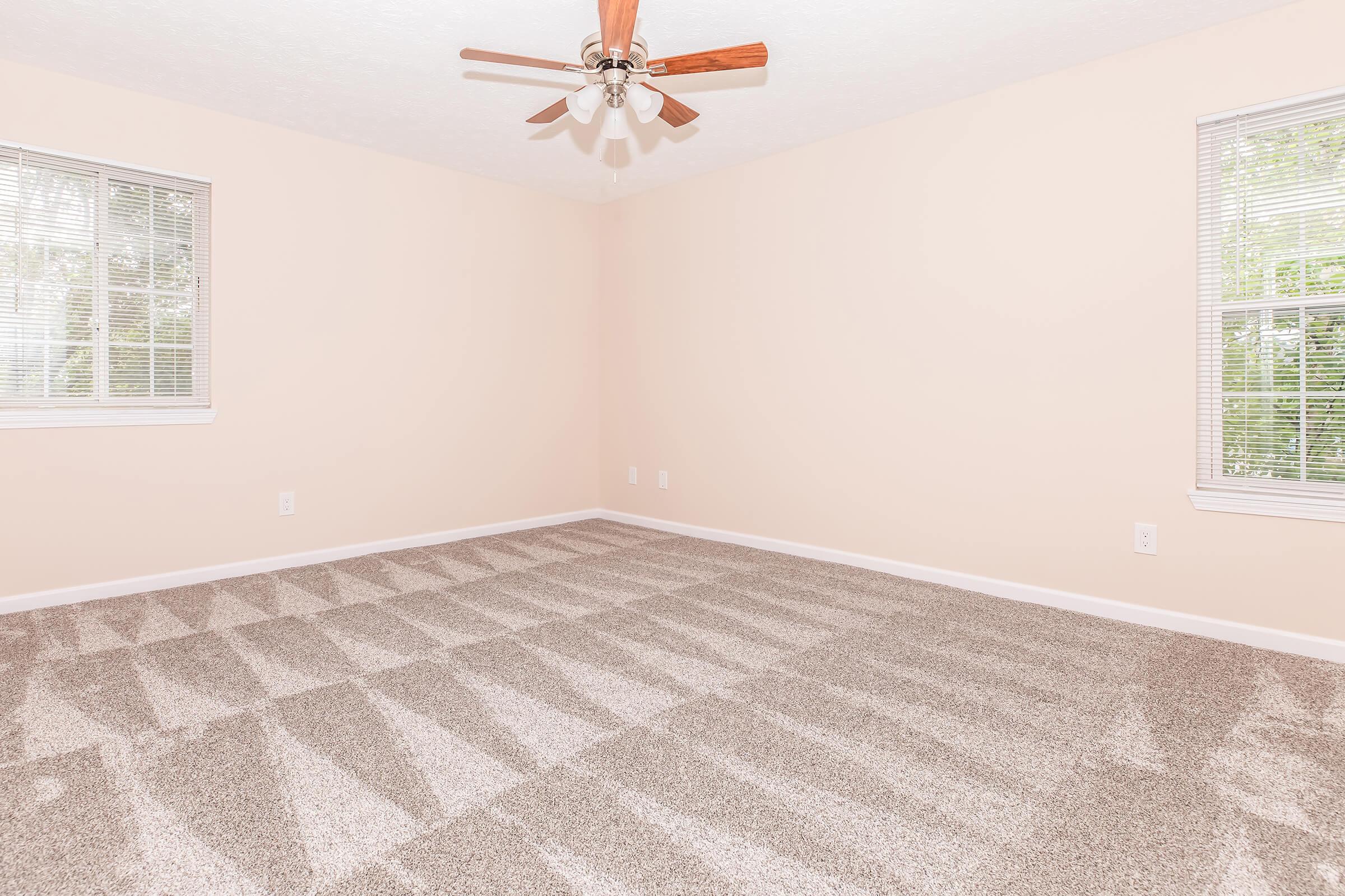 Empty room with light beige walls and light brown carpet. Two windows on the left side allowing natural light, and a ceiling fan with wooden blades in the center. The floor shows a pattern created by light and shadow, enhancing the spacious feel of the area.