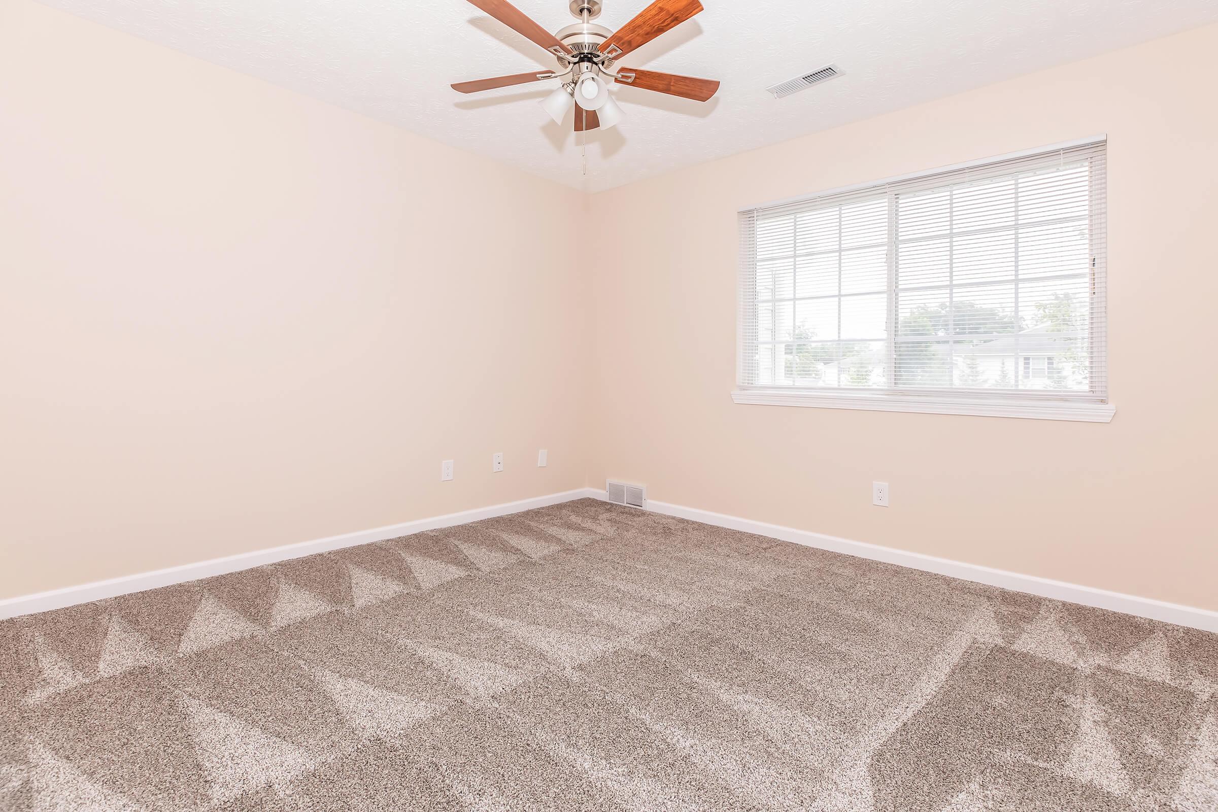 A spacious, empty bedroom with light beige walls and a large window letting in natural light. The ceiling features a ceiling fan, and the floor is covered in plush, light brown carpet with a geometric pattern.