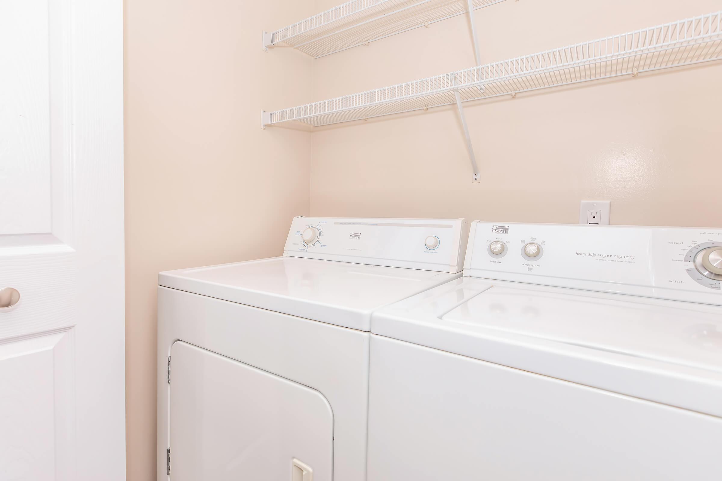 A clean laundry room featuring a white washer and dryer side by side. The walls are painted a soft beige, and there are wire shelves mounted above the appliances for storage. A white door is partially visible, adding to the organized and tidy appearance of the space.