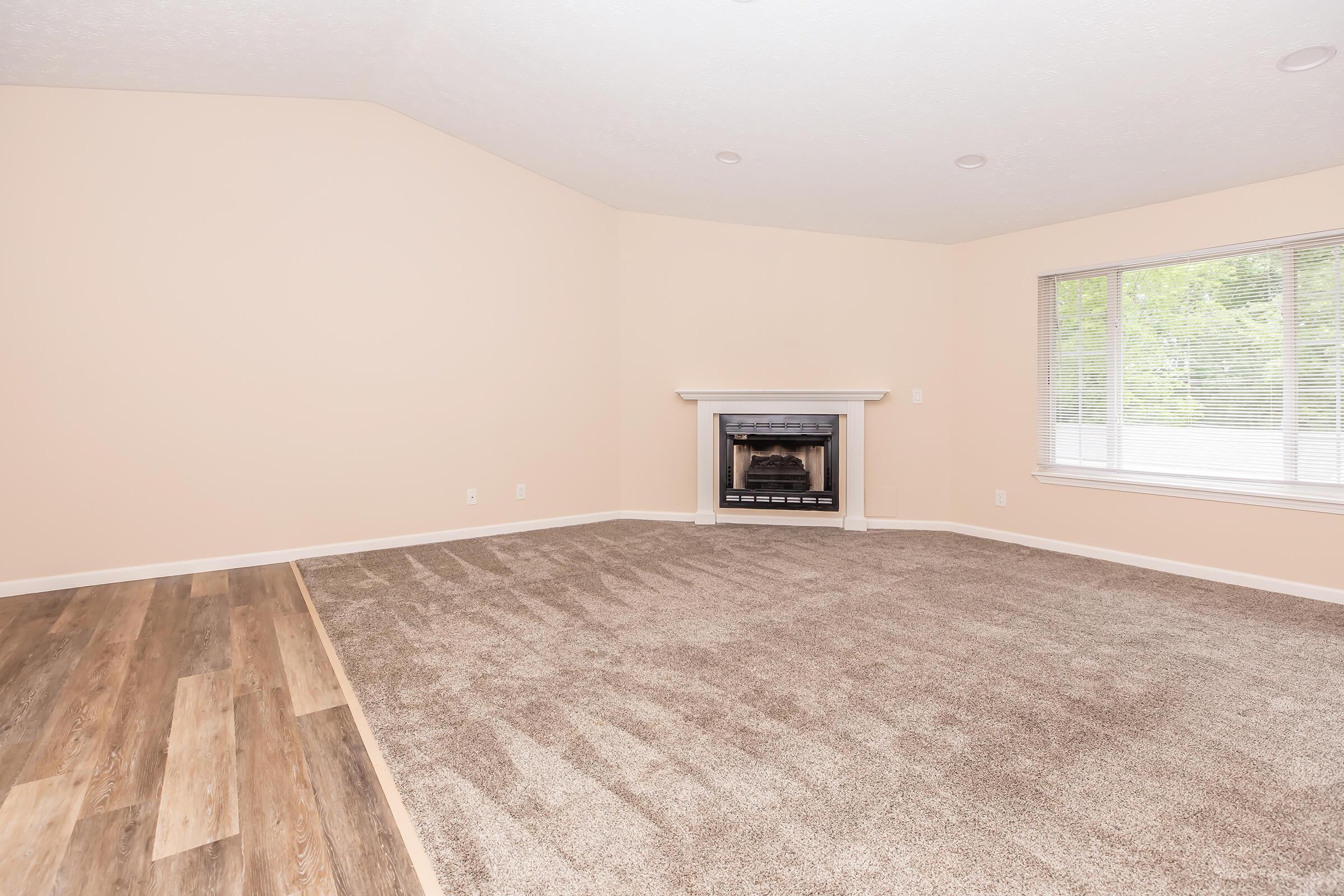 A spacious, softly lit living room with light beige walls and a large window allowing natural light. The floor features a mix of carpet and hardwood, and there is a fireplace with a white mantel against one wall, creating a cozy focal point.