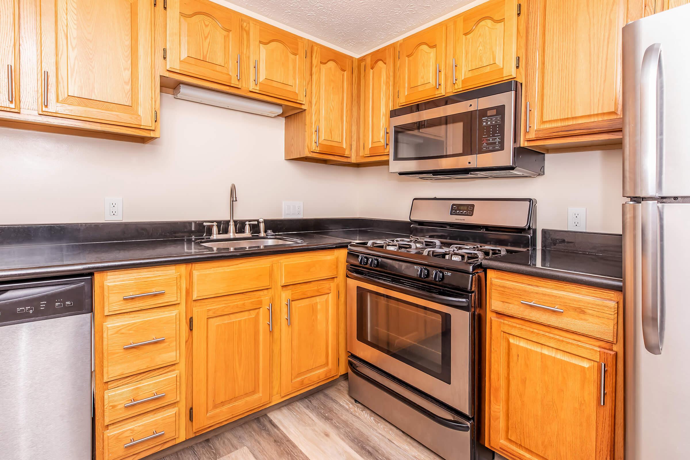 Modern kitchen featuring wooden cabinets, stainless steel appliances including a microwave and oven, black countertops, and a dishwasher. The space is well-lit and organized, showcasing a functional layout for cooking and meal preparation.