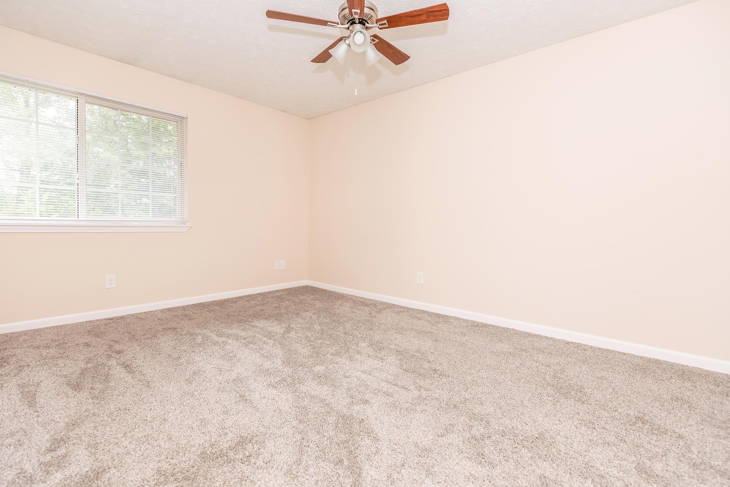 A vacant room with beige walls and light gray carpet. It features a ceiling fan with wooden blades, a large window with natural light coming in, and no furniture. The space appears clean and ready for occupancy.
