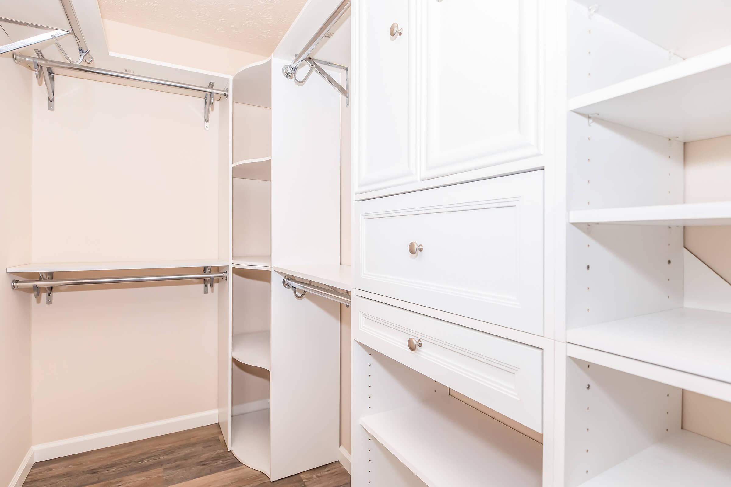 A spacious, organized closet featuring white cabinetry, open shelves, and a hanging rod. The design includes drawers for storage and a neutral wall color, creating an inviting and functional space for clothing and accessories.