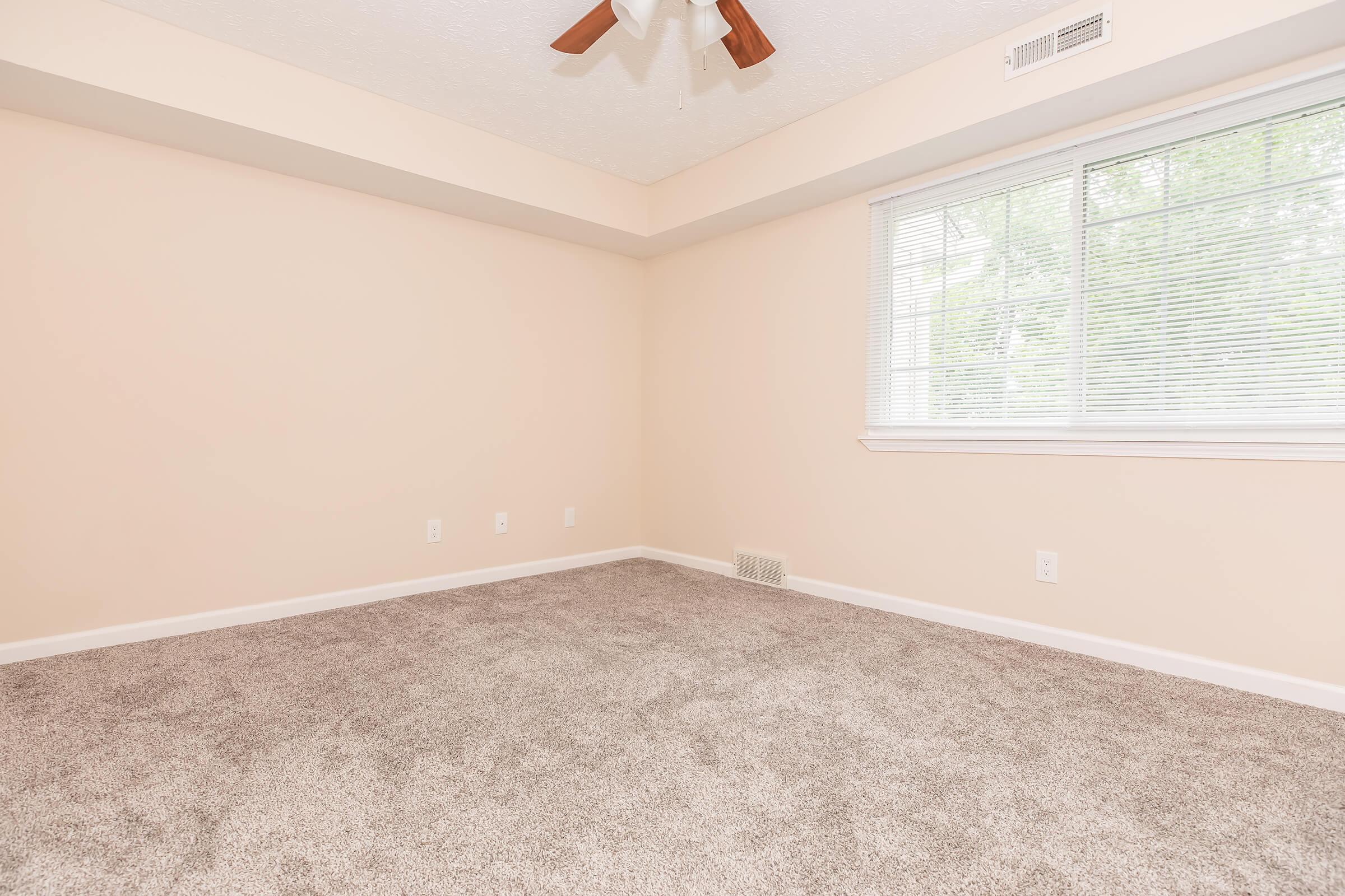 A spacious, empty room with light peach walls and a plush carpet. There is a ceiling fan with wooden blades, and a large window allowing natural light to fill the space. The overall atmosphere is bright and inviting, perfect for personalization or decorating.