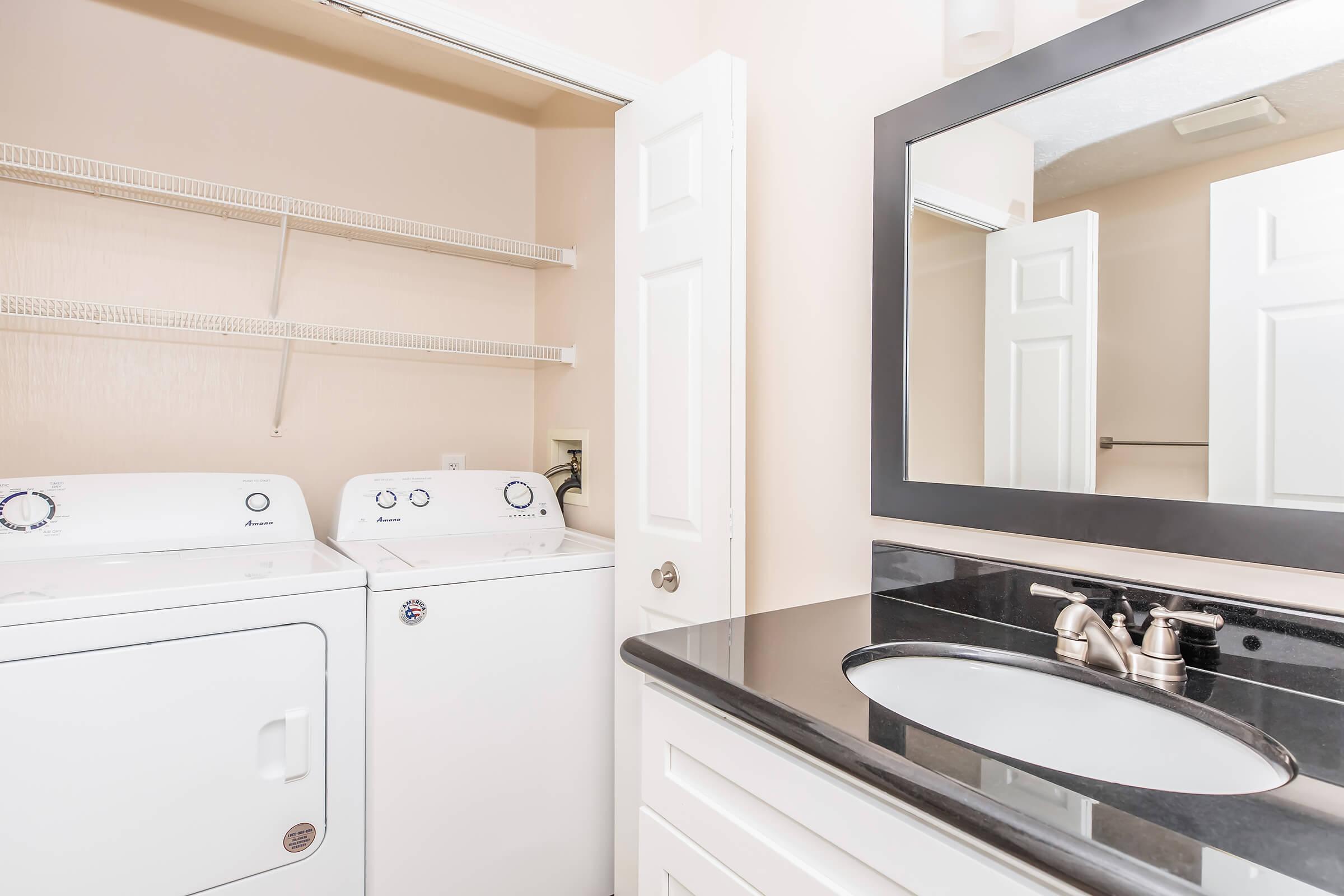 A laundry room featuring a stacked washer and dryer on the left, with shelves above. To the right, there is a modern bathroom vanity with a black granite countertop and a round sink, complemented by a large mirror above it. The walls are painted in a light color, creating a clean and organized space.