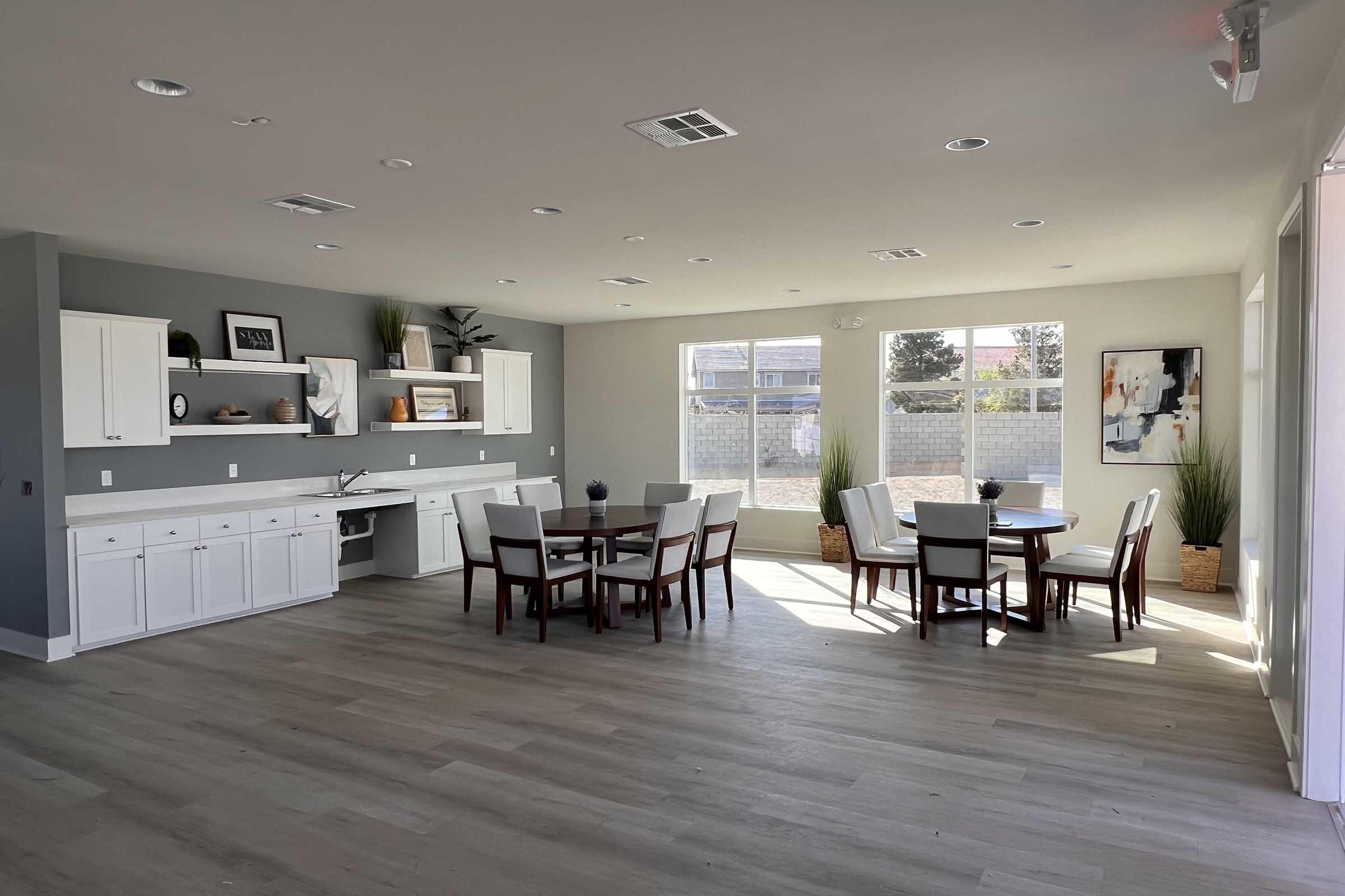Bright and modern interior of a spacious room featuring a dining area with a round table and chairs, white cabinetry, and shelves displaying decorative items. Large windows allow natural light to fill the space, complemented by plants in woven baskets and neutral-colored walls and flooring.