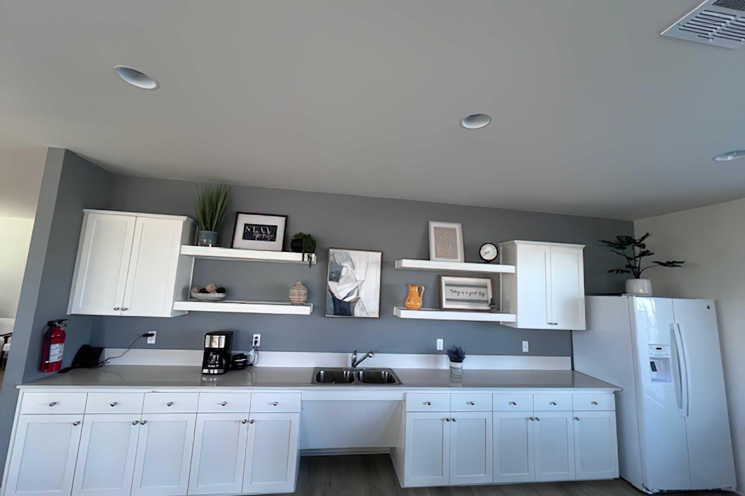 A modern kitchen featuring white cabinets and open shelving. The gray walls complement a minimalist design that includes decorative items, a coffee maker, and a white refrigerator. Natural light enhances the space, which has a clean and organized appearance.