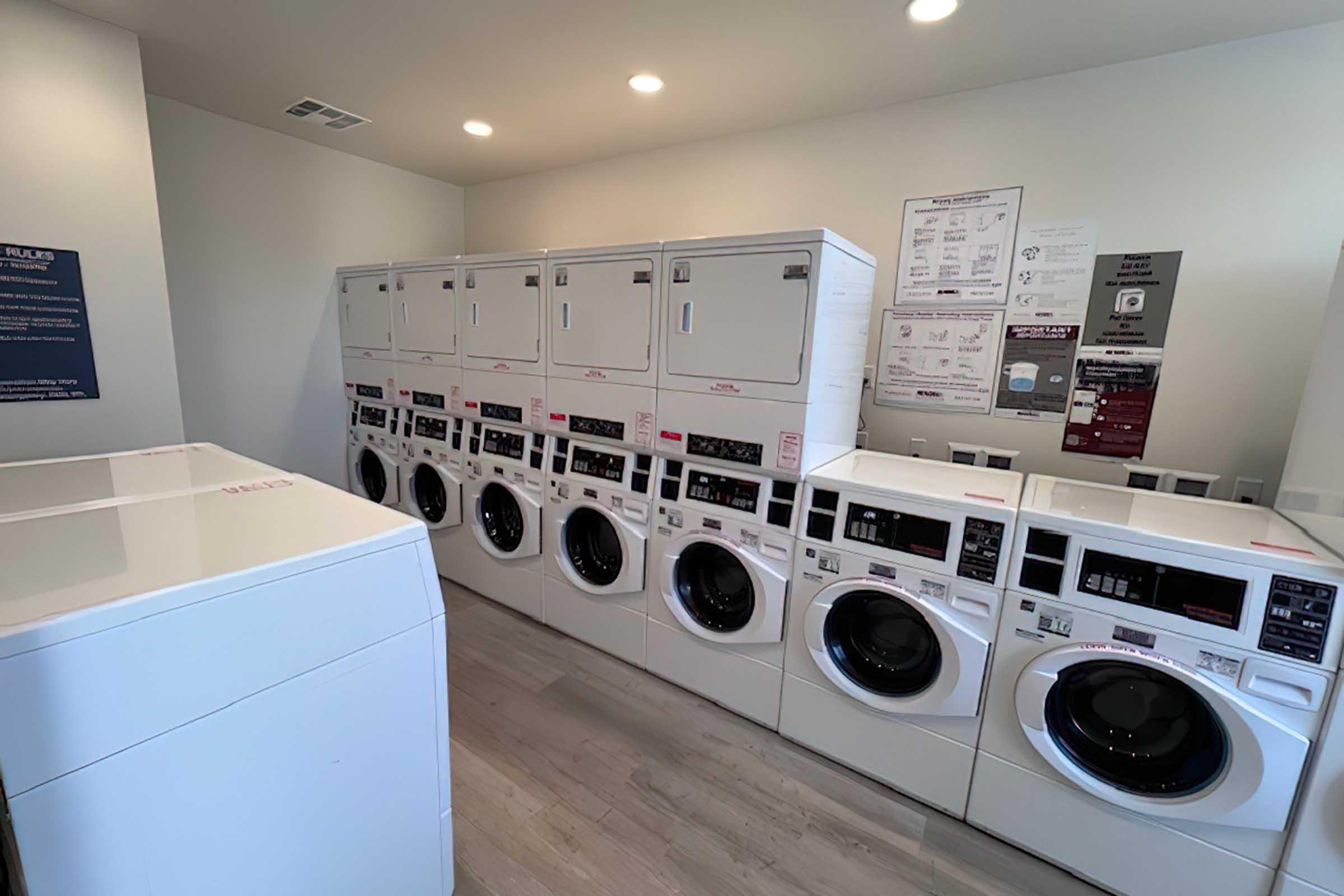 A laundry room featuring multiple washing machines and dryers lined up against the wall. The machines are modern and white, with instructions posted nearby. The floor is light-colored wood, and the room has bright lighting, creating a clean and functional environment for laundry services.