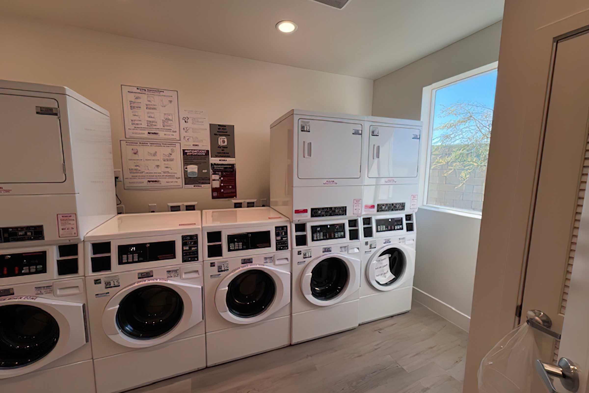 A bright laundry room featuring multiple stacked and side-by-side washing machines and dryers. White appliances are neatly arranged along the wall, with informational posters displayed nearby. A window allows natural light to enter, illuminating the space with a view of trees outside.