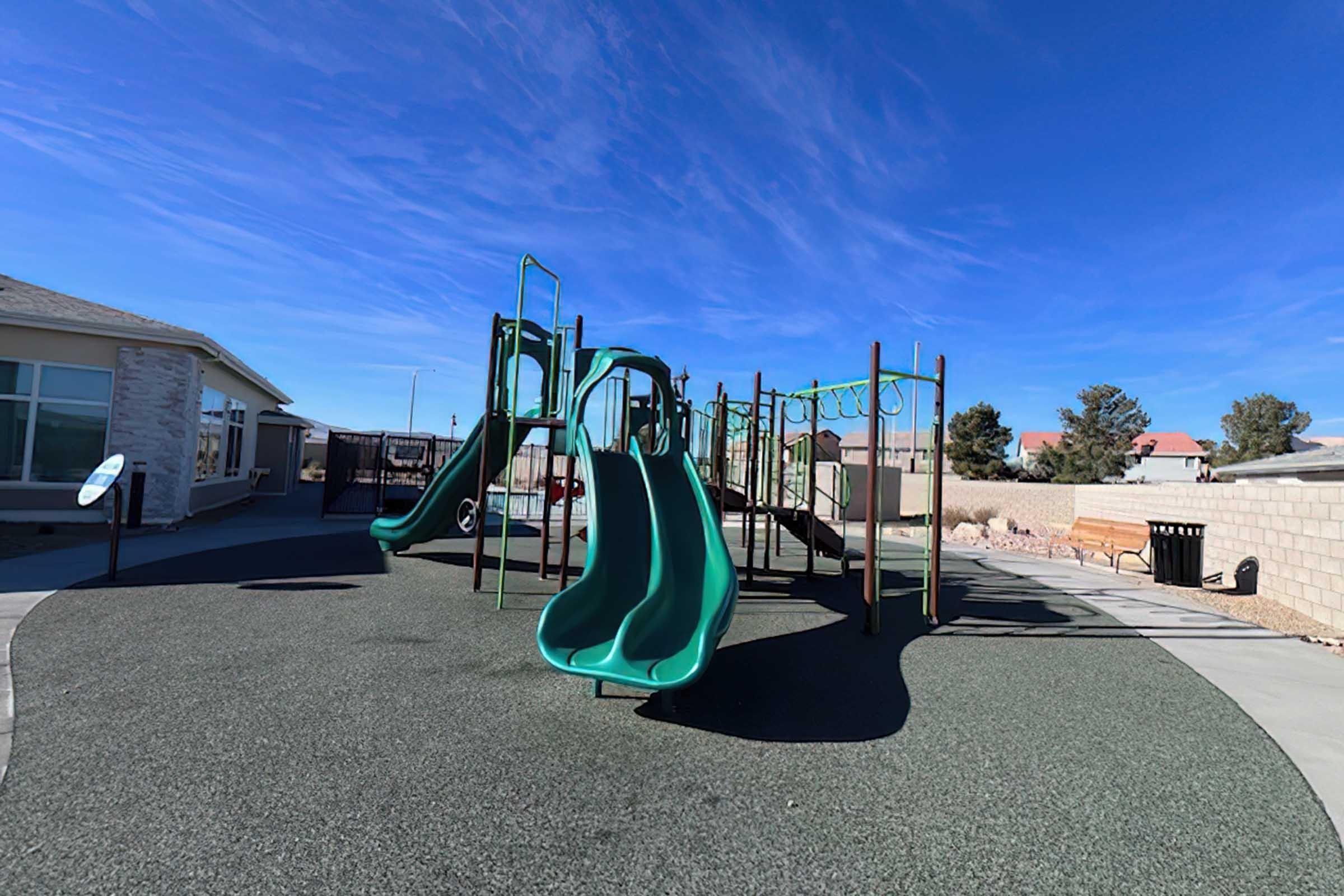 A playground featuring two green slides, a climbing structure, and a safety surface made of rubber and gravel. In the background, there is a clear blue sky and a residential building, with surrounding landscaping and a fence.
