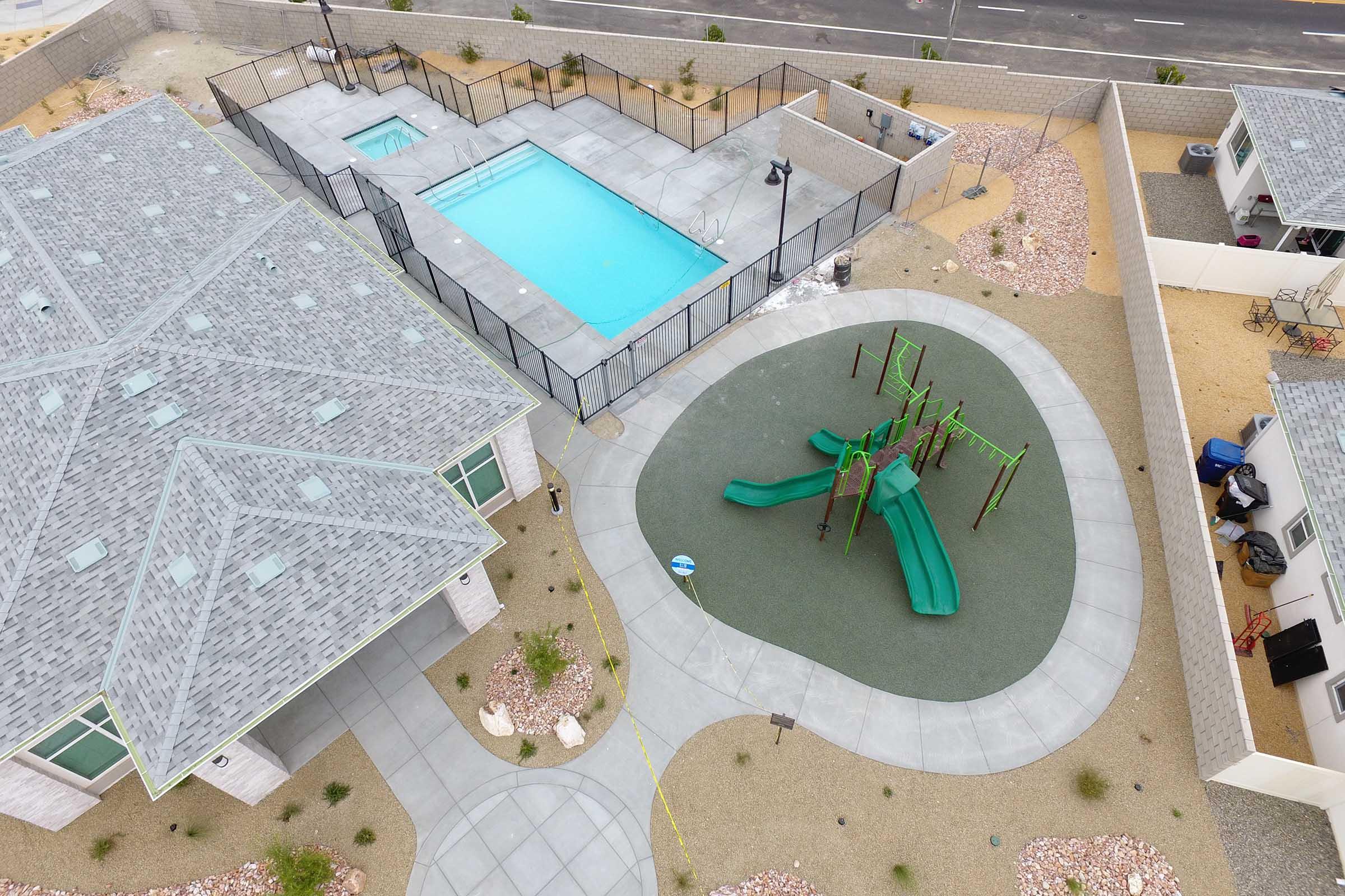 An aerial view of a residential area featuring a fenced swimming pool and a playground. The playground includes green slides and climbing structures, surrounded by gravel landscaping and pathways. Nearby, there are houses with gray roofs and a clear view of the road in the background.