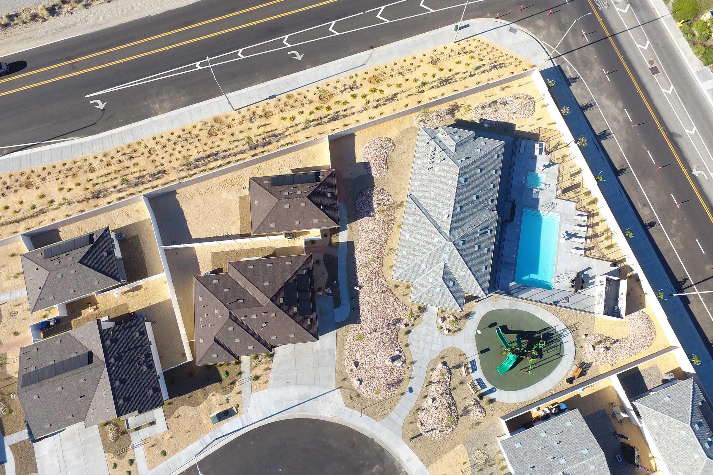 Aerial view of a residential neighborhood featuring multiple houses with brown roofs, landscaped yards, and a community pool area with green play equipment. Surrounding roads and pathways are visible, along with desert landscaping and plants.