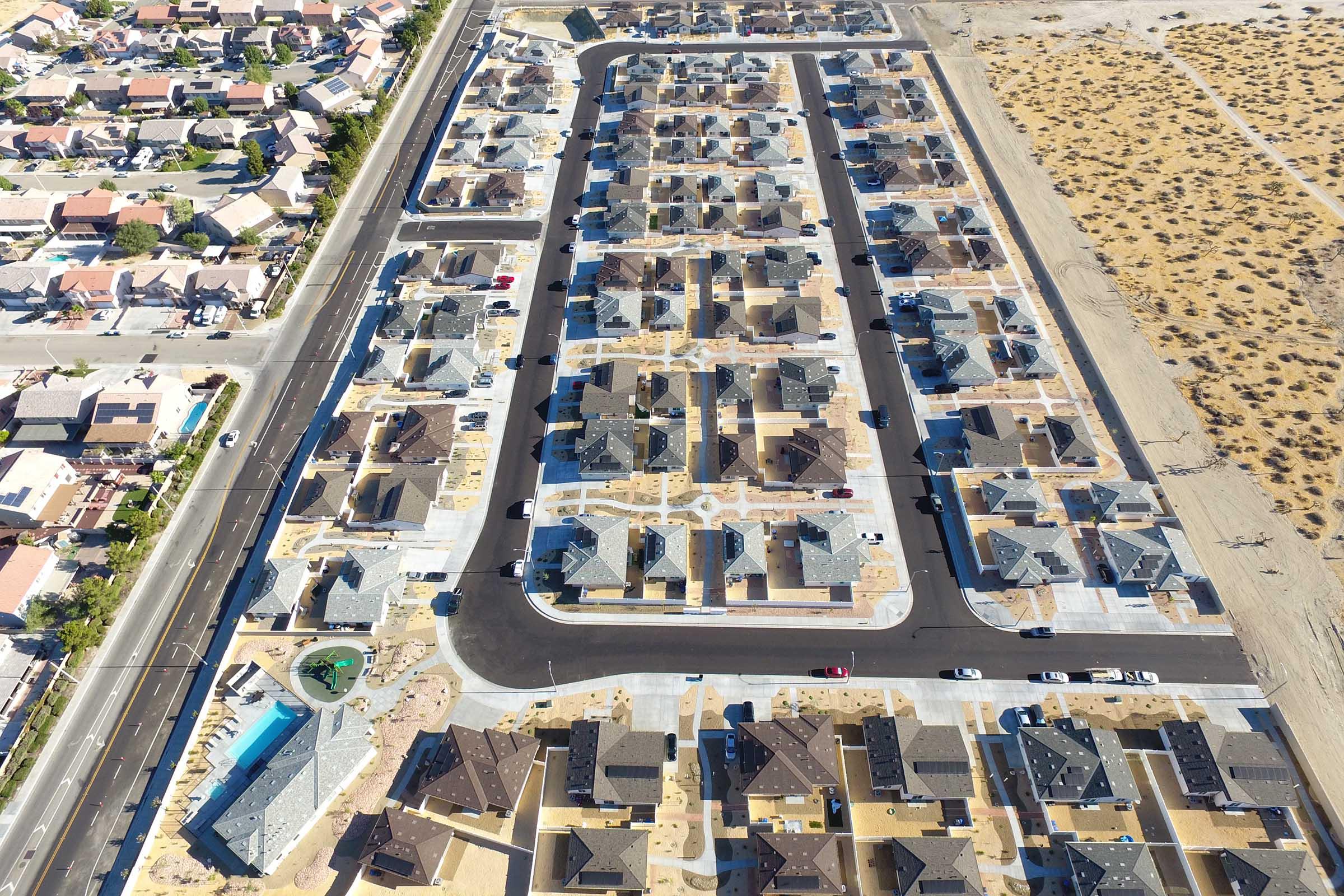 Aerial view of a residential neighborhood featuring numerous houses arranged in a grid pattern, with a nearby road and some sparse desert landscape. The houses have dark roofs and are surrounded by empty lots, indicating a recently developed area.