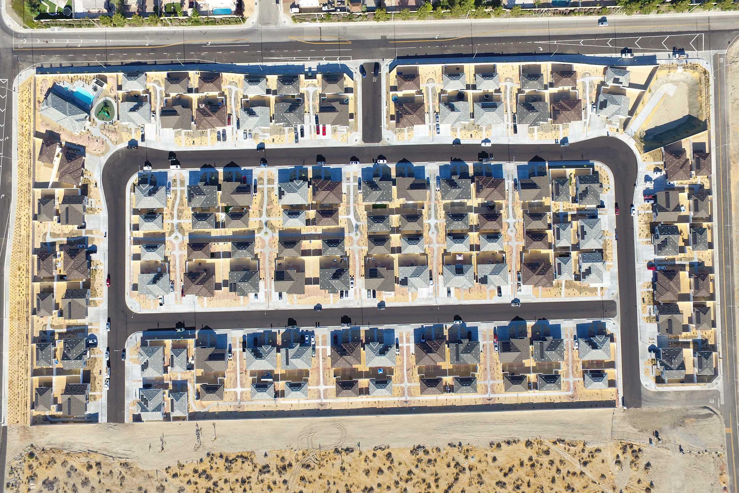 Aerial view of a neighborhood featuring a grid layout of houses, arranged in rows with driveways and landscaped yards. The area is mostly cleared of vegetation, with some new construction visible. The streets are paved, and shadows from the buildings create geometric patterns on the ground.