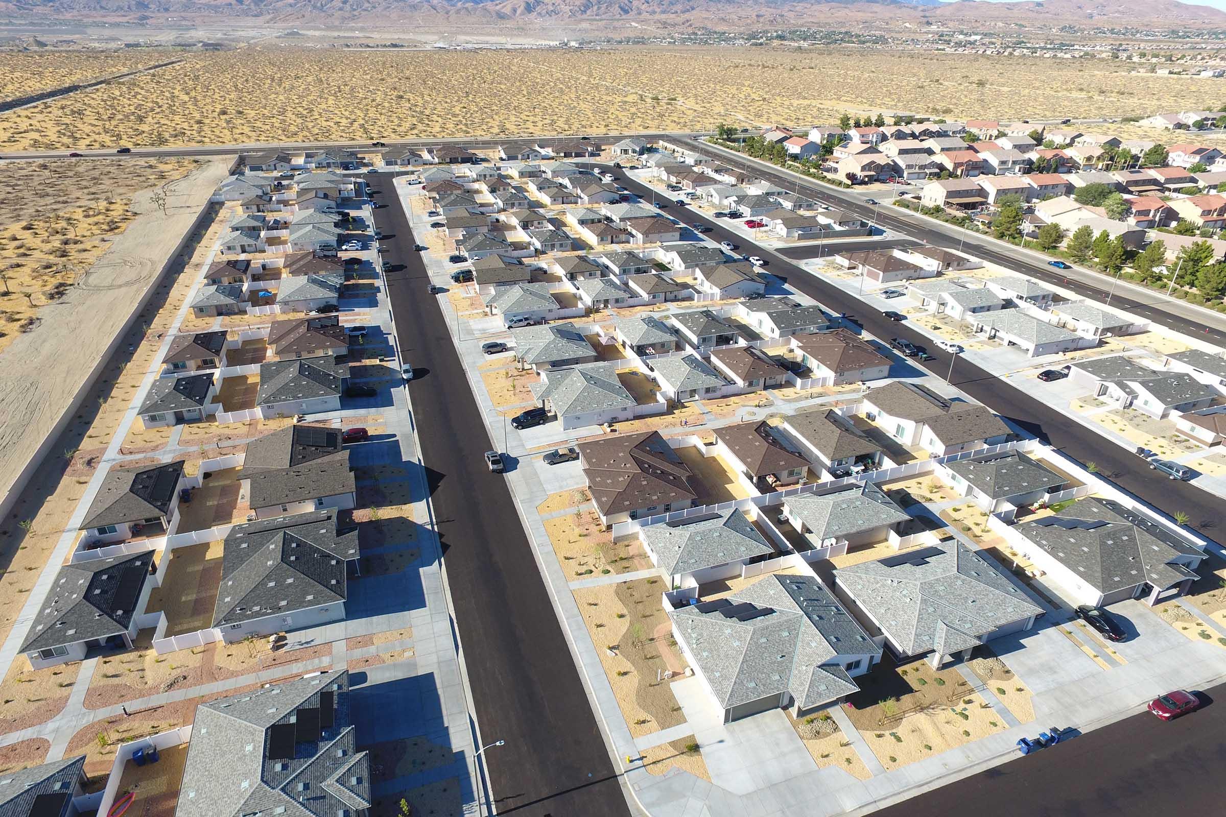 Aerial view of a suburban neighborhood featuring rows of single-family homes with a variety of roof styles. The area is surrounded by open land and mountains in the background, with some vehicles parked on the streets. The landscape shows arid terrain with minimal landscaping around the houses.