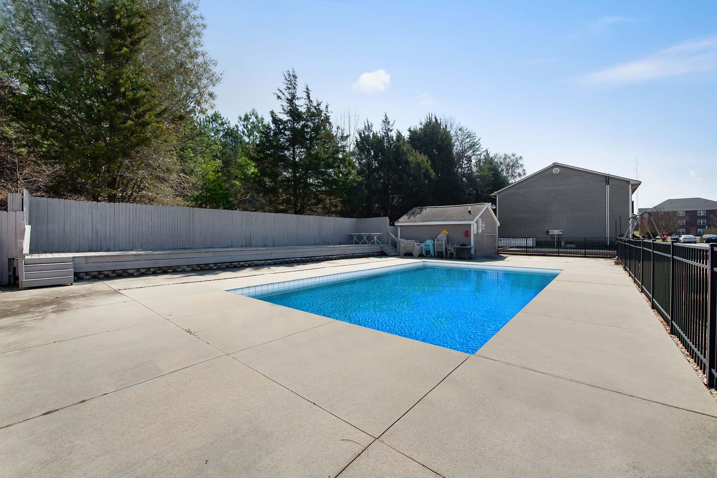 A clear blue swimming pool surrounded by a concrete deck. In the background, there are tall trees and a gray building. There are also chairs and a storage shed near the pool area, with a fence enclosing the space. The sky is bright and blue with a few clouds.