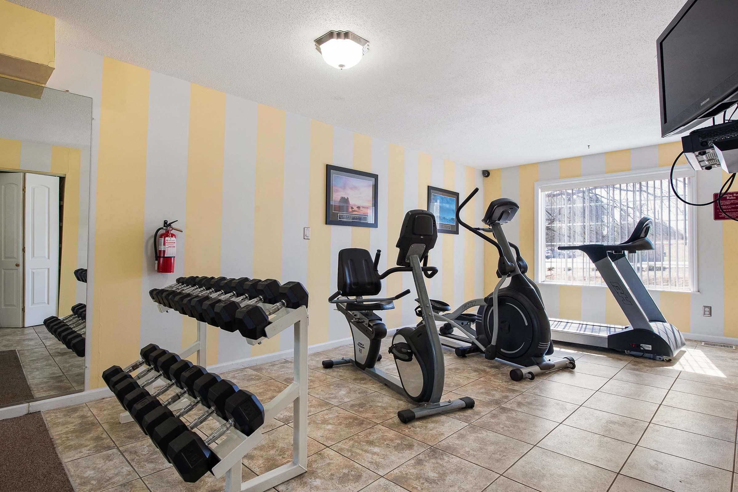 A fitness room featuring exercise equipment, including a stationary bike, elliptical machine, and a rack of dumbbells. The walls are painted in yellow and white stripes, and there’s a large mirror and a fire extinguisher visible. Large windows provide natural light.