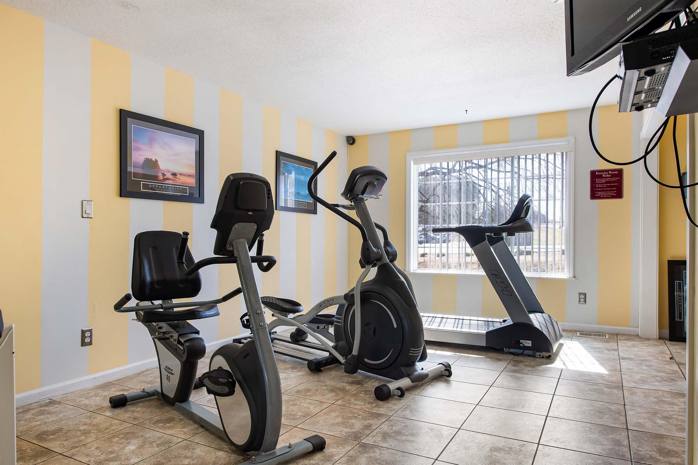 A small fitness room featuring two exercise bikes, a treadmill, and a television. The walls are painted with yellow and white stripes, and there are framed pictures on the wall. A window provides natural light, and there's a notice on the wall nearby. The floor is tiled.