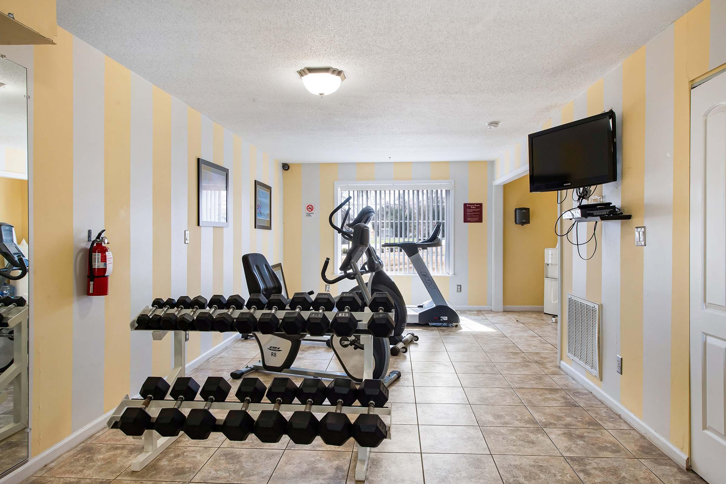 A small fitness room featuring a set of dumbbells, an elliptical machine, a flat-screen TV mounted on the wall, and a fire extinguisher. The walls are painted with yellow and white stripes, and there are windows providing natural light. The space has a tiled floor.