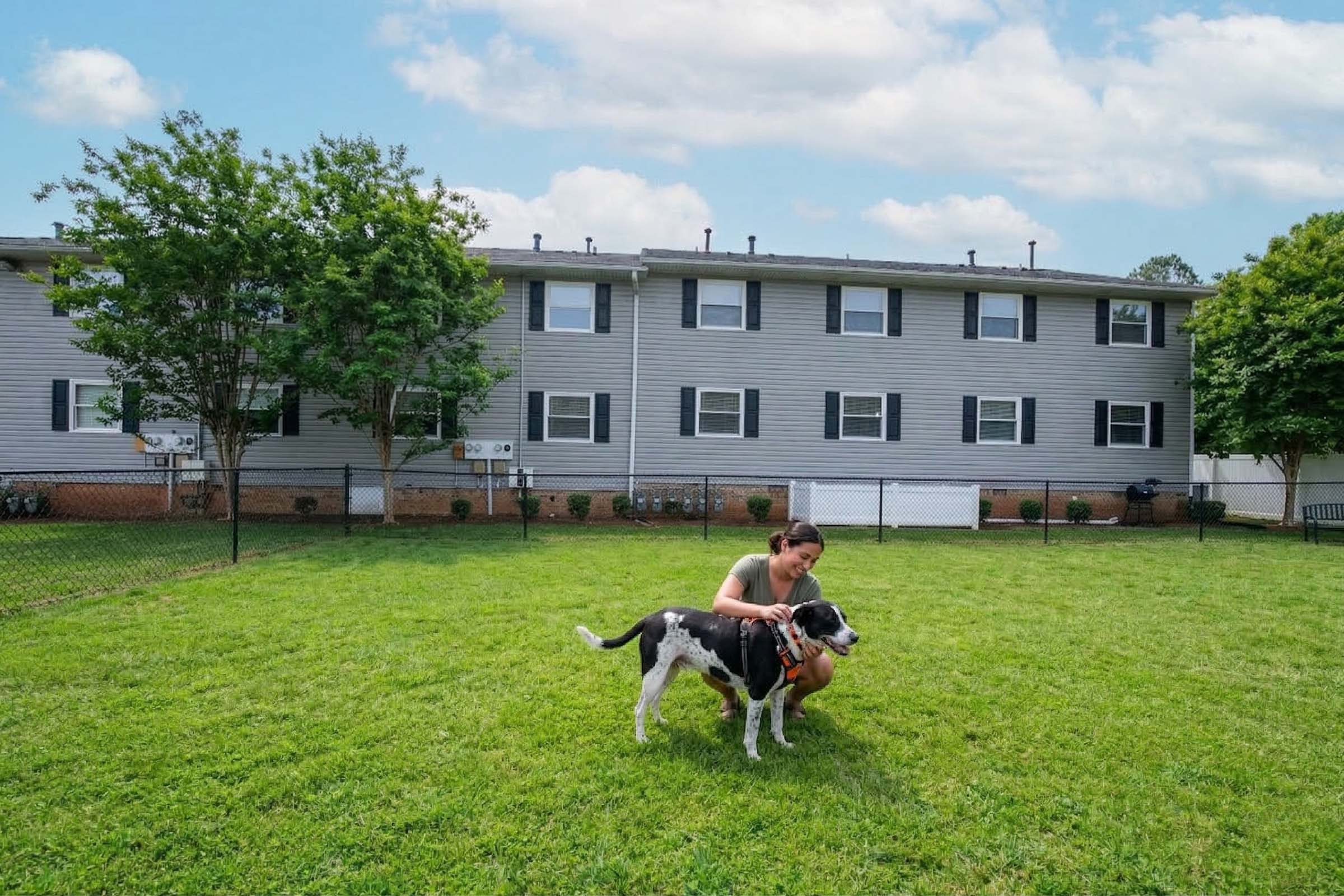 A woman kneels on a lush green lawn, interacting with a black and white dog. In the background, there are several apartment buildings with multiple windows and greenery around them. The sky is partly cloudy, creating a bright, pleasant atmosphere.