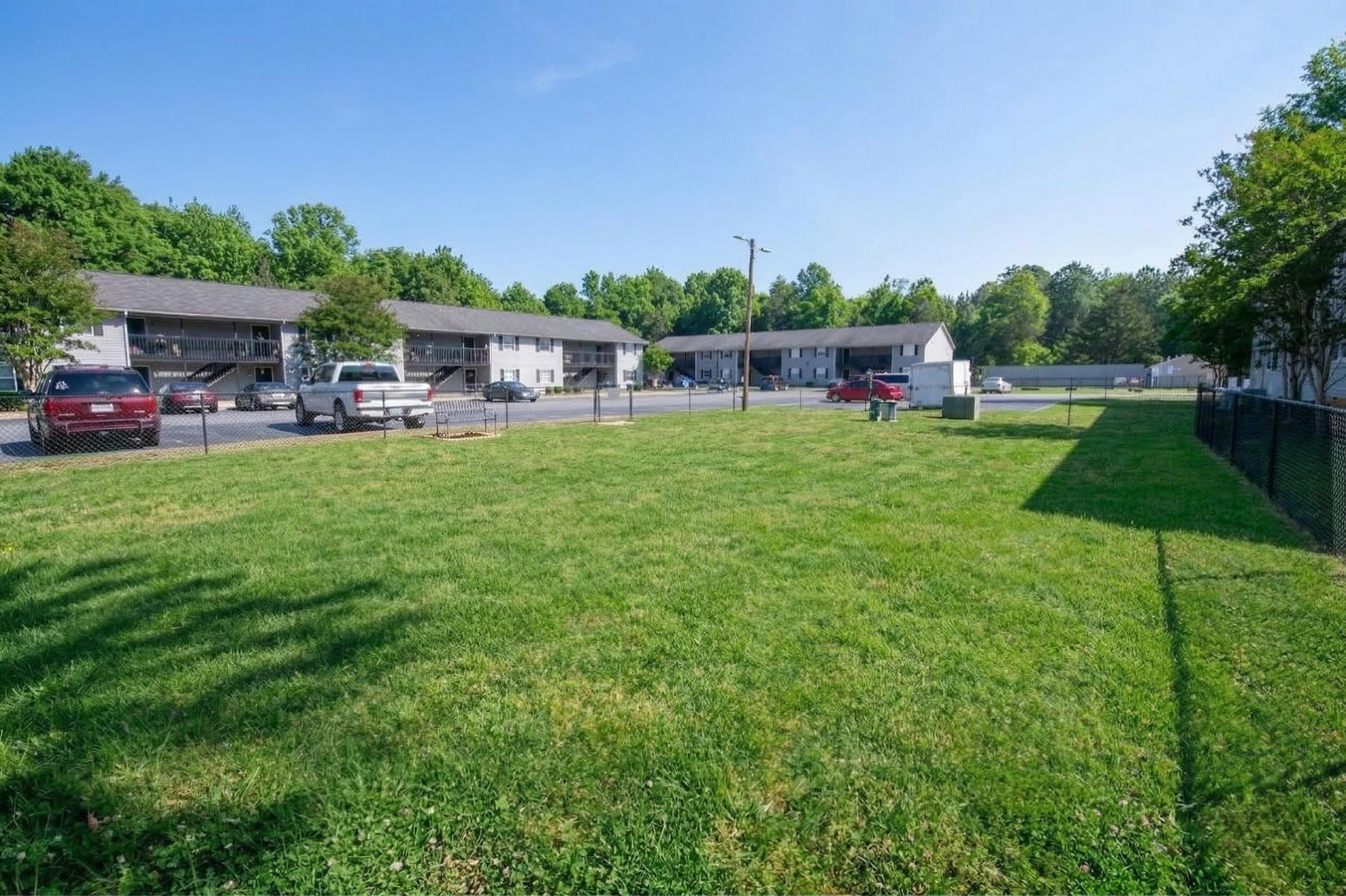 A grassy area in a residential neighborhood with several multi-family buildings in the background. There are parked cars in the lot, and a fence surrounding the grassy space. The scene is set on a clear day with blue skies and greenery in the surrounding area.