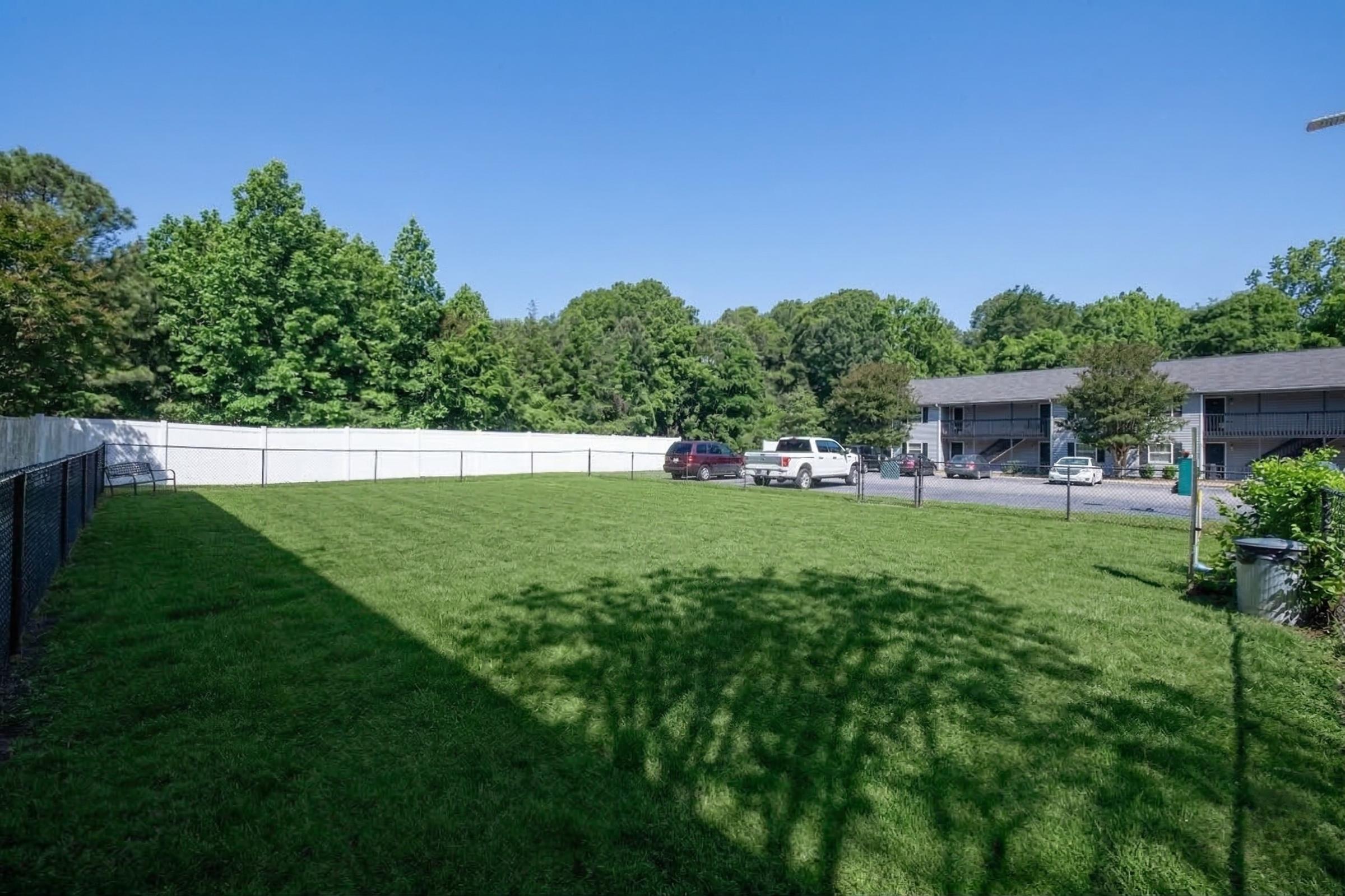 A grassy area enclosed by a fence, featuring several trees and a building in the background. Two parked cars are visible on a driveway. The scene is bathed in sunlight on a clear day, emphasizing the lush greenery and well-maintained space.