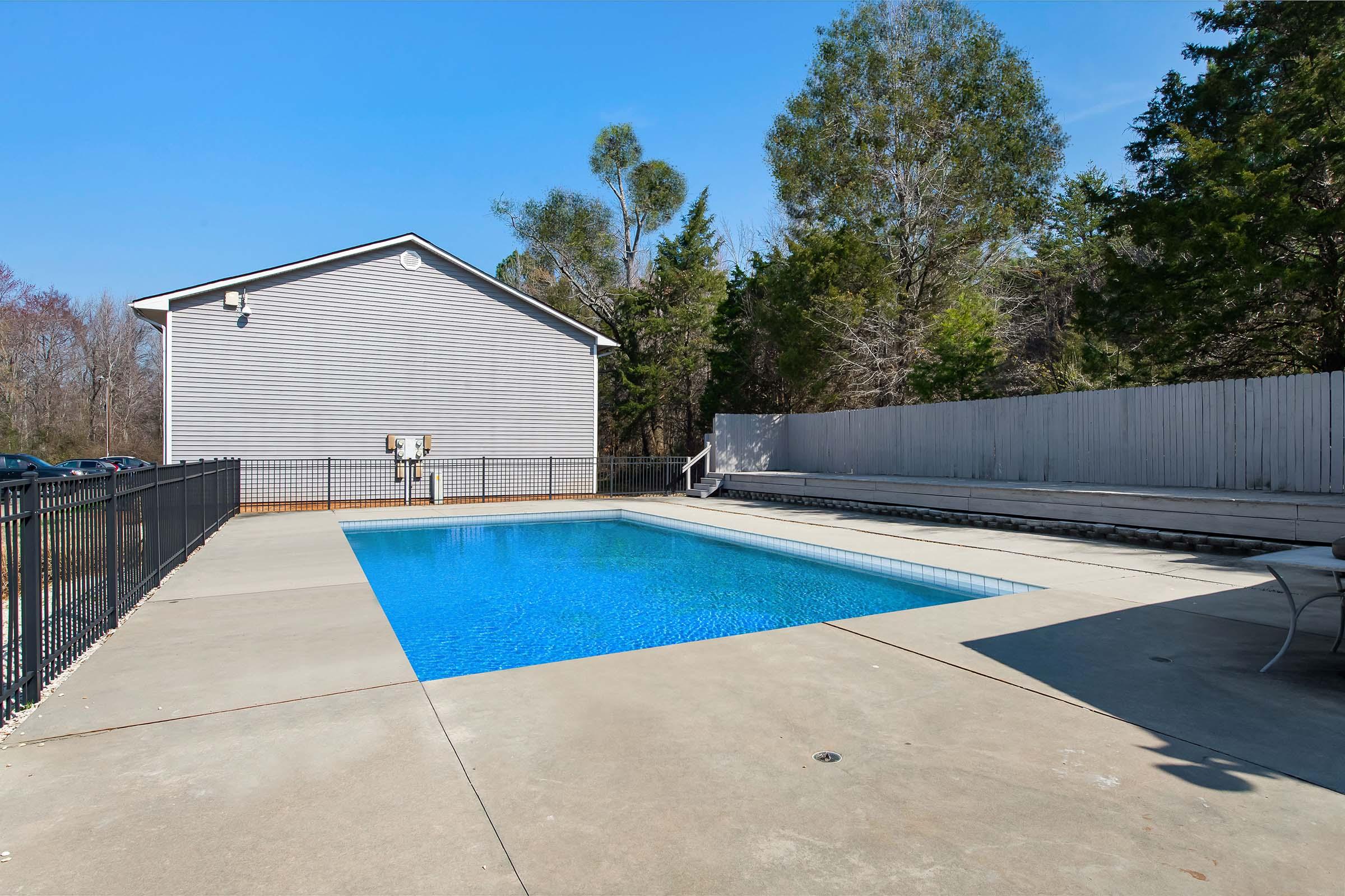 A clean, blue swimming pool surrounded by a concrete patio and a black metal fence. Lush greenery and trees can be seen in the background, along with a light gray building. The sky is clear and blue, indicating a sunny day.