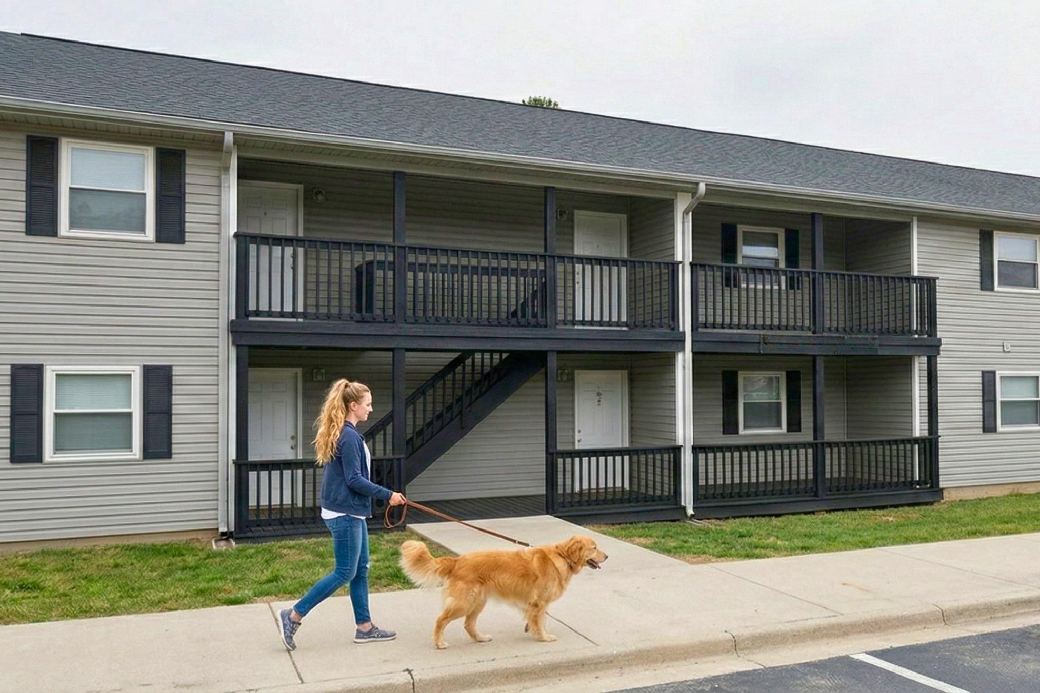 A young woman walks a golden retriever on a leash in front of a two-story apartment building. The building has a light gray exterior with black railings on the balconies. The woman wears a blue sweater and jeans, and there are two doors visible on the ground level of the building.