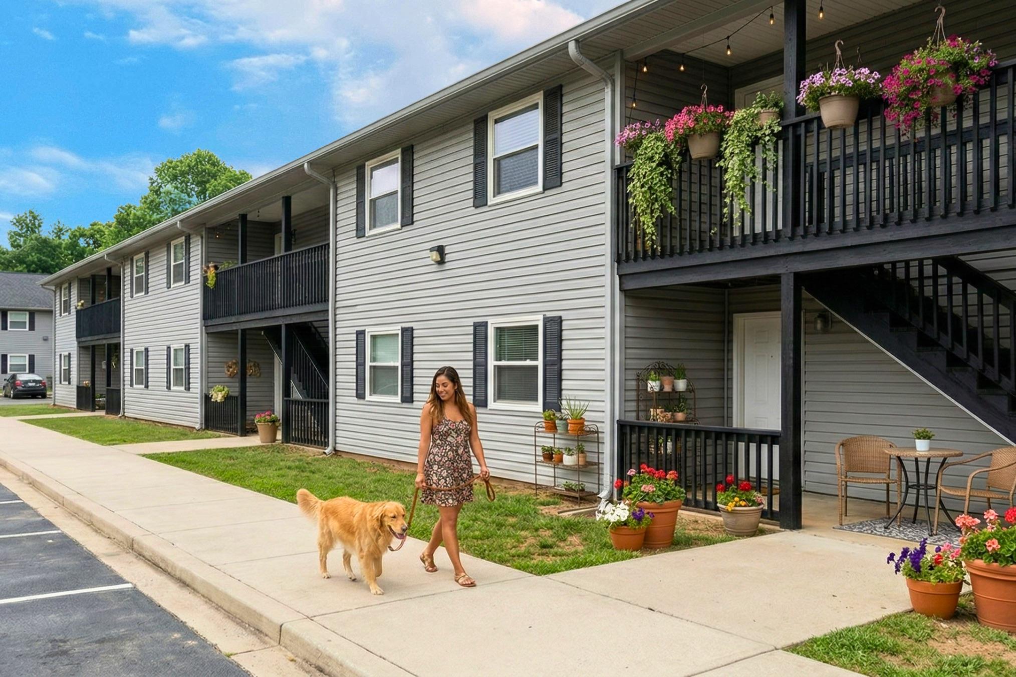A woman in a floral dress walks a golden retriever along a sidewalk in front of a two-story apartment building. The building features decorative plants and flowers on the balconies, and there are chairs and potted plants near the entrance. The sky is partly cloudy, creating a bright and welcoming atmosphere.