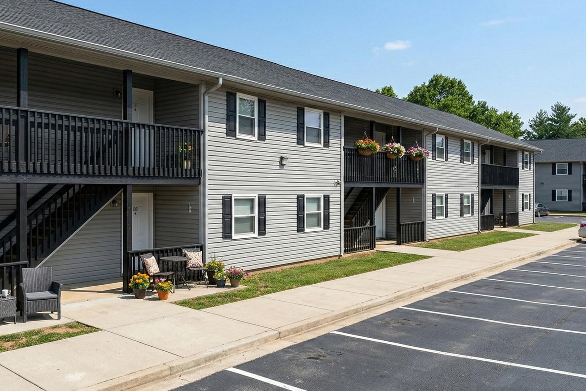 Two-story apartment building with grey siding and black railings. Balconies adorned with flower pots. Sidewalk leading to the entrance, with seating areas and parking spaces in front. A landscaped area with grass and trees in the background under a clear blue sky.