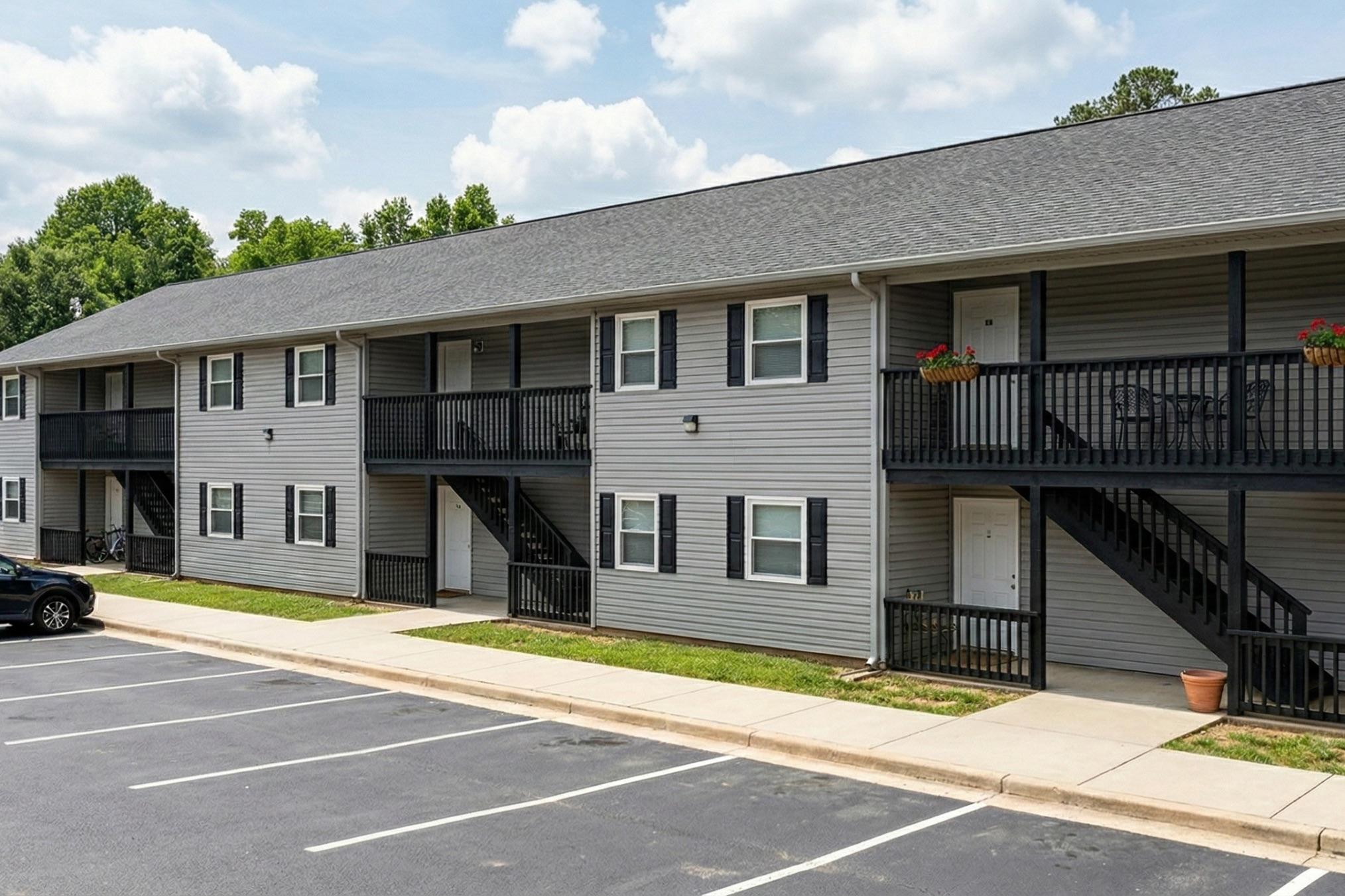 A low-rise apartment building featuring multiple units with gray siding. Each unit has a balcony and a staircase. The building is surrounded by a grassy area and a paved parking lot with several empty spaces. The sky is partly cloudy.