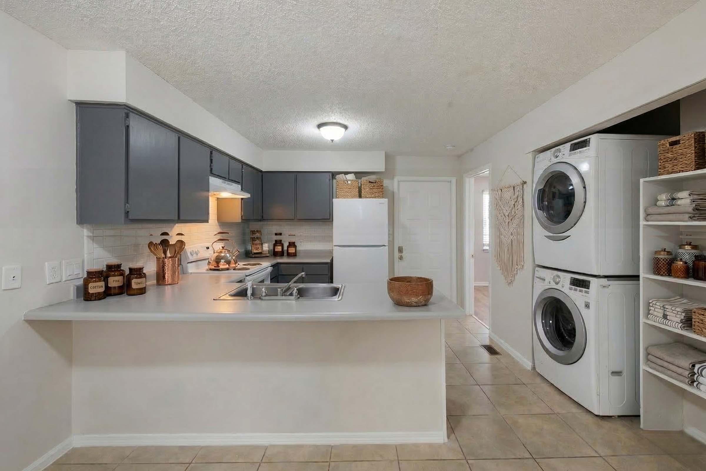 A modern kitchen featuring gray cabinets, a double sink, and a refrigerator. Adjacent to the kitchen is a laundry area with a stacked washer and dryer. The space includes decorative containers on shelves and a warm light fixture, creating an inviting atmosphere. The flooring is tiled for easy maintenance.
