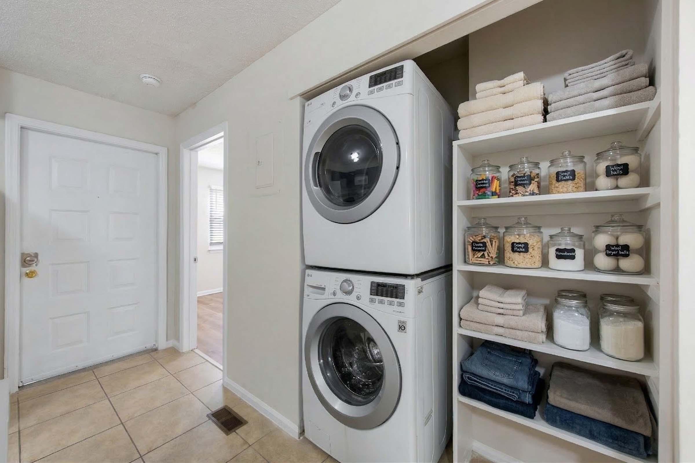 A compact laundry area featuring a stacked washer and dryer beside a shelf. The shelf is neatly organized with jars containing various laundry supplies and labeled containers, along with folded towels and blankets. The wall is painted in a light color, and there is a door leading to another room in the background.