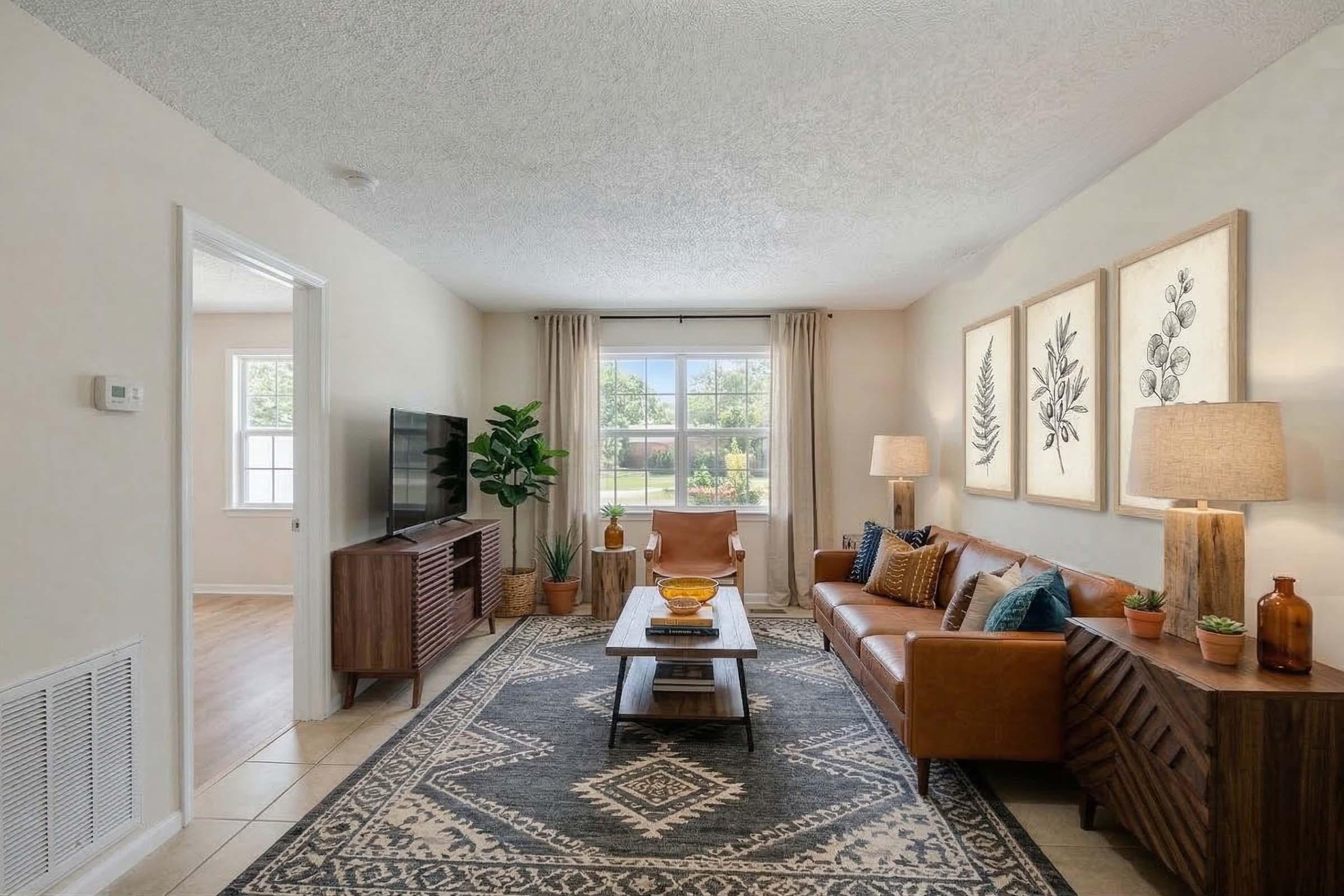A cozy living room featuring a brown leather sofa adorned with decorative pillows, a wooden coffee table with a bowl, and a patterned area rug. There are two stylish lamps on either side, potted plants, and framed botanical artwork on the wall. A large window lets in natural light, illuminating the space.