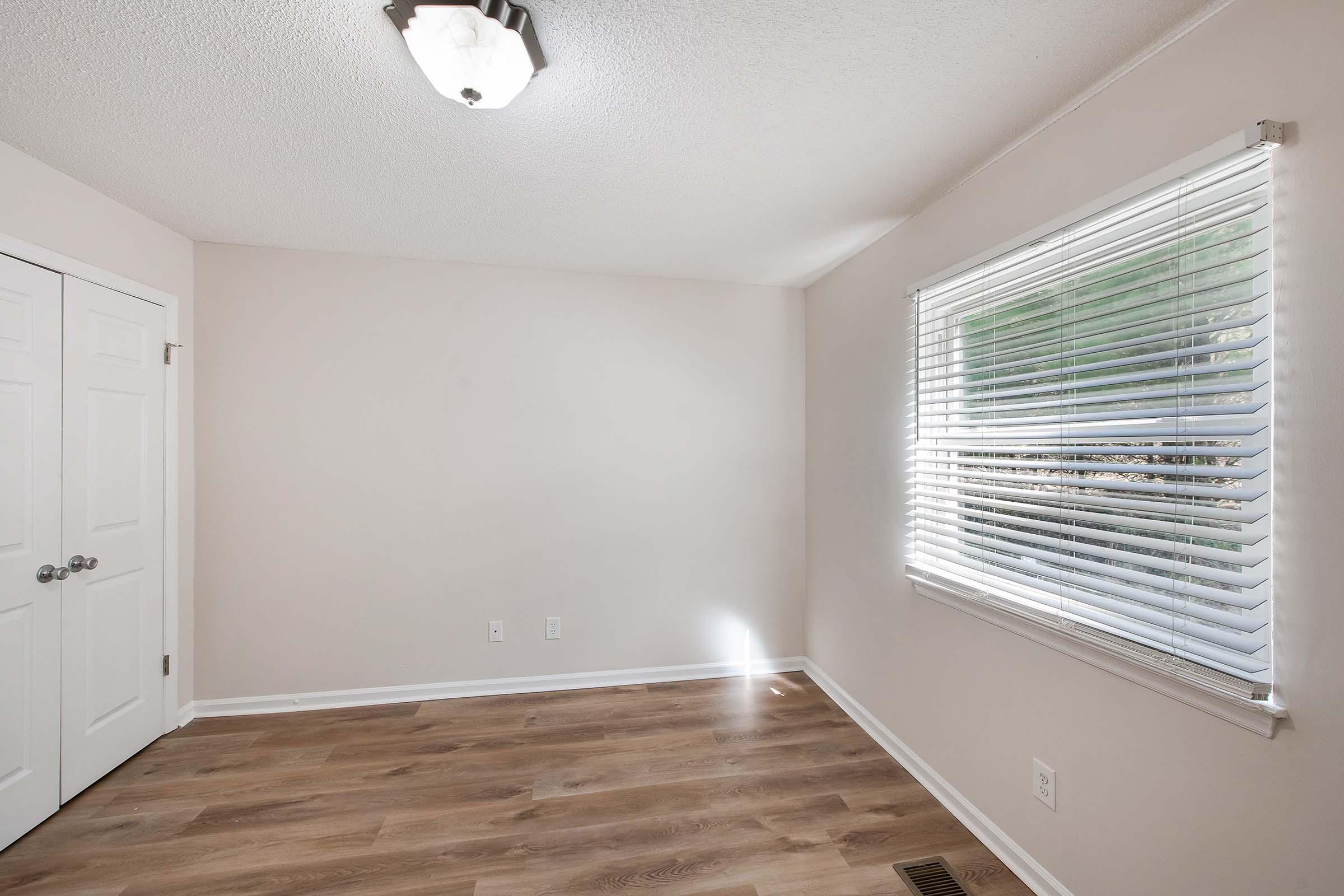 Empty room with light-colored walls and wooden flooring. A window with white horizontal blinds allows natural light to enter. The ceiling features a simple light fixture, and there are two closed white doors on one side of the room.