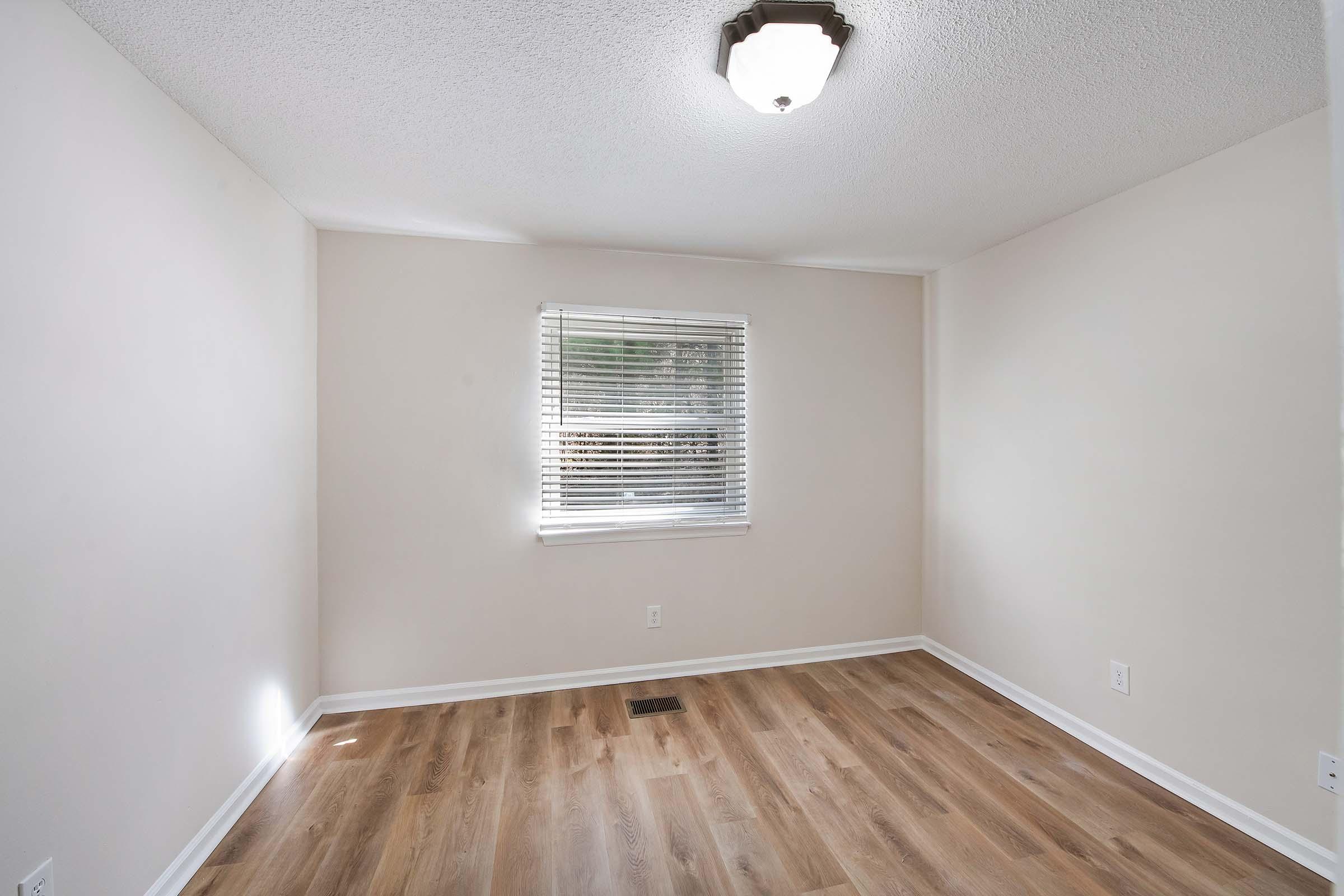 A bright, empty room with light-colored walls and a wooden floor. A window with white blinds allows natural light to enter, and there is a ceiling light fixture. The space appears clean and freshly painted, suitable for various uses like a bedroom or office.