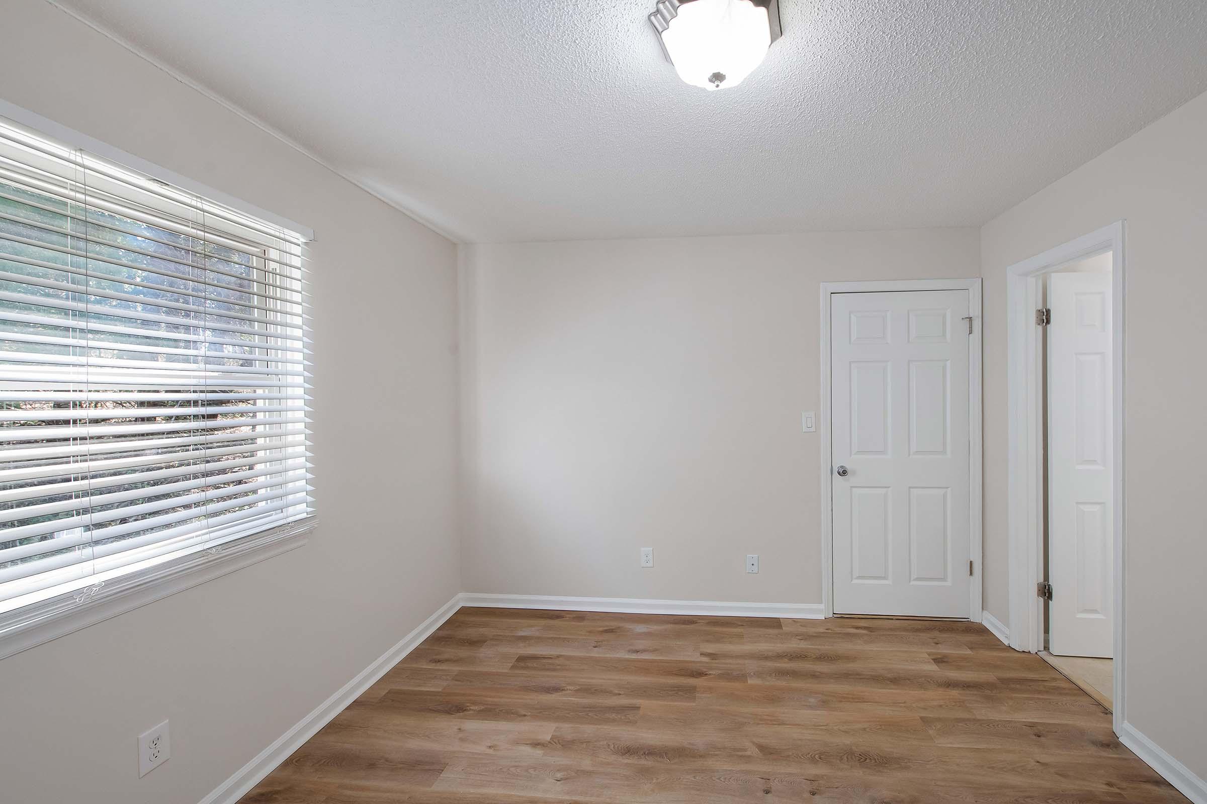A vacant room featuring light-colored walls, laminate flooring, and a ceiling light fixture. Natural light enters through a window with blinds. There are two doors, one leading to a closet and the other to an adjacent space. The room has a clean and minimalistic appearance, suitable for various uses.