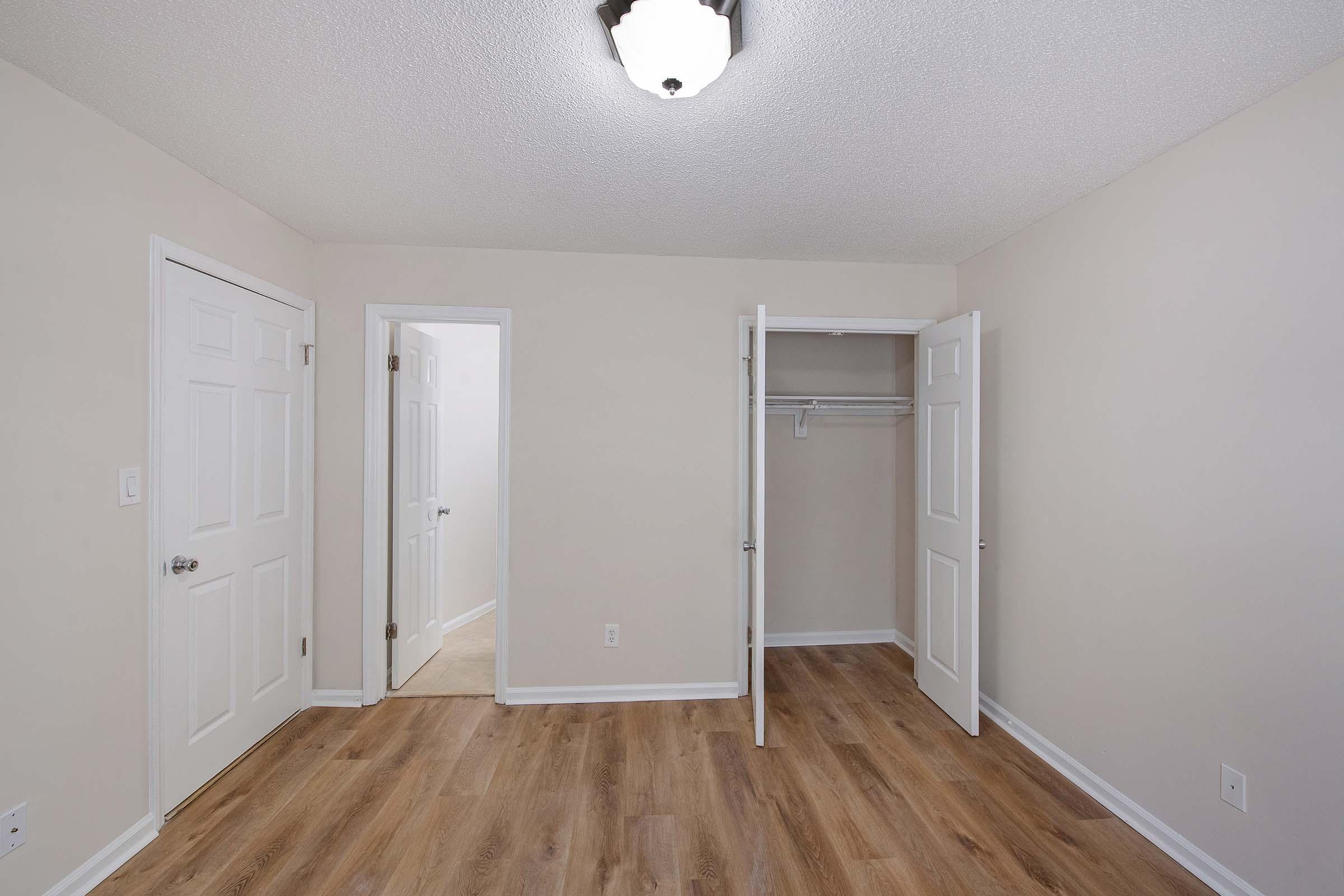 A spacious, empty room with beige walls and wooden floors. There are two closed doors on the left, one leading to another room and the other to a closet with open doors. A ceiling light fixture is visible, illuminating the area. The room is well-lit and free of furniture, creating a blank canvas for decoration.