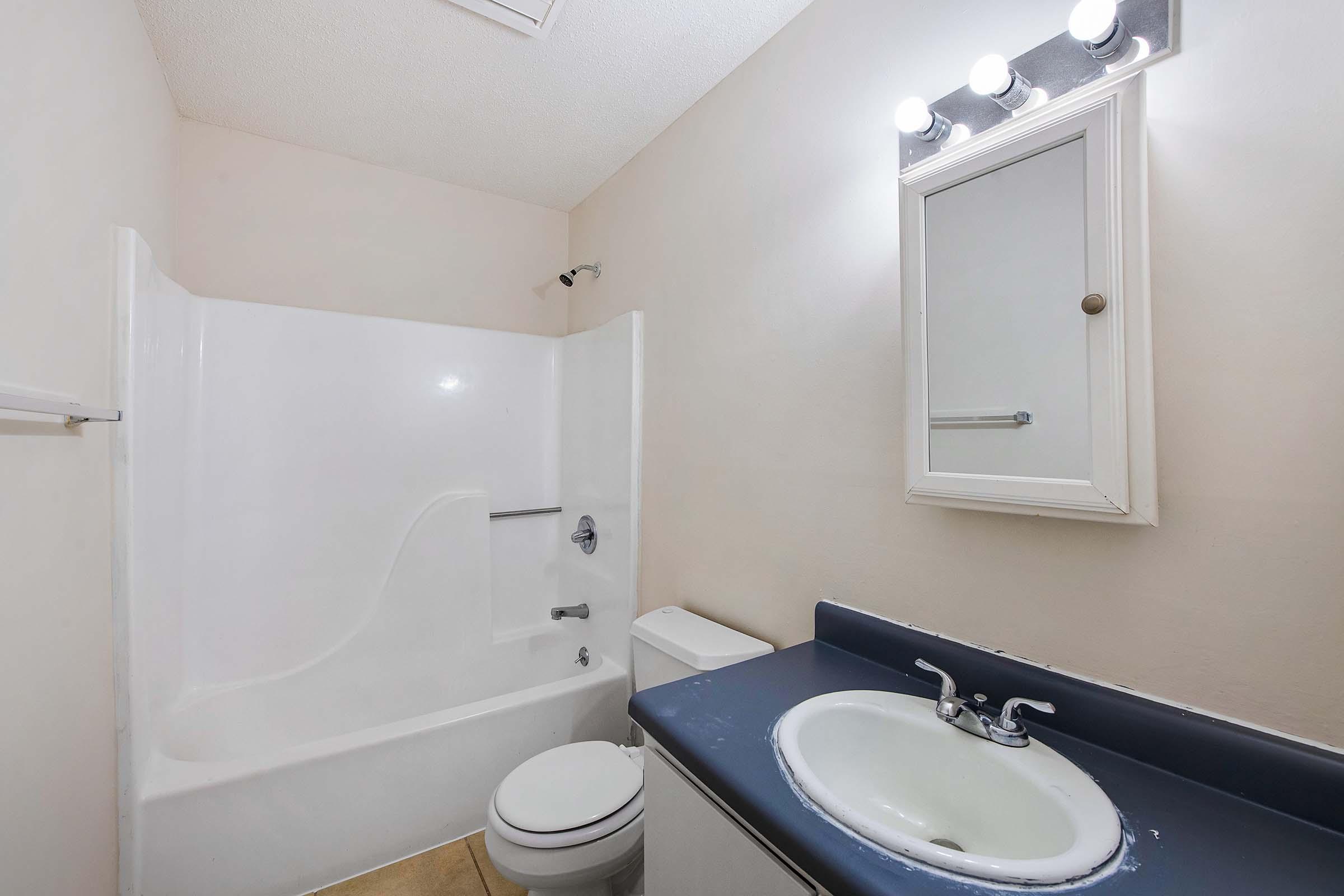A small bathroom featuring a white bathtub with a shower, a toilet, and a sink with a blue countertop. There is a mirror above the sink and a light fixture with three bulbs. The walls are painted in a light color, and the flooring is tiled.