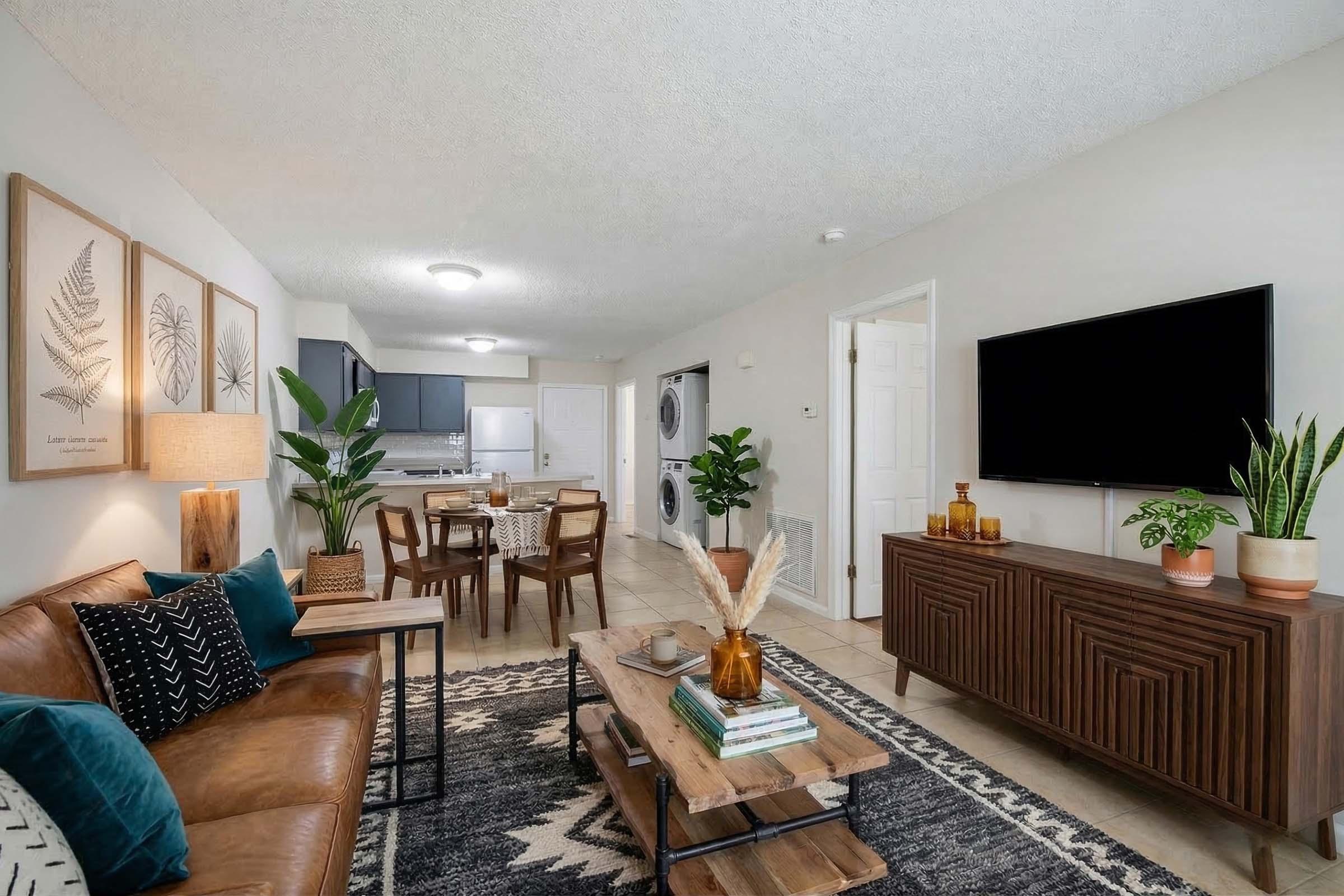 A modern living room featuring a brown leather sofa adorned with teal and white pillows, a coffee table with books and decorative items, and potted plants. The walls display framed botanical prints, and there's a dining area in the background with a round table and chairs. A large TV is mounted on the wall.