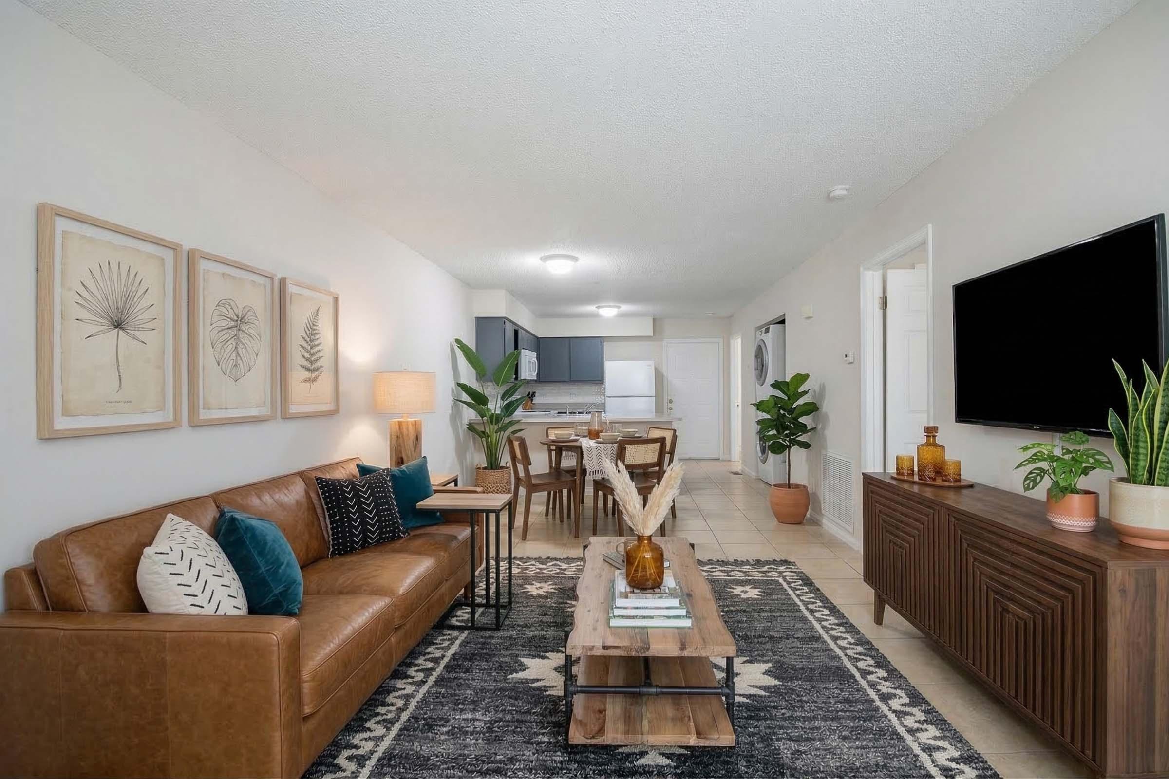 A modern living room with a brown leather sofa adorned with cushions and a coffee table made of wood. There are framed botanical prints on the wall, a dining area with a wooden table and chairs, and a kitchen visible in the background. Indoor plants add a touch of greenery, with a television mounted on the wall.