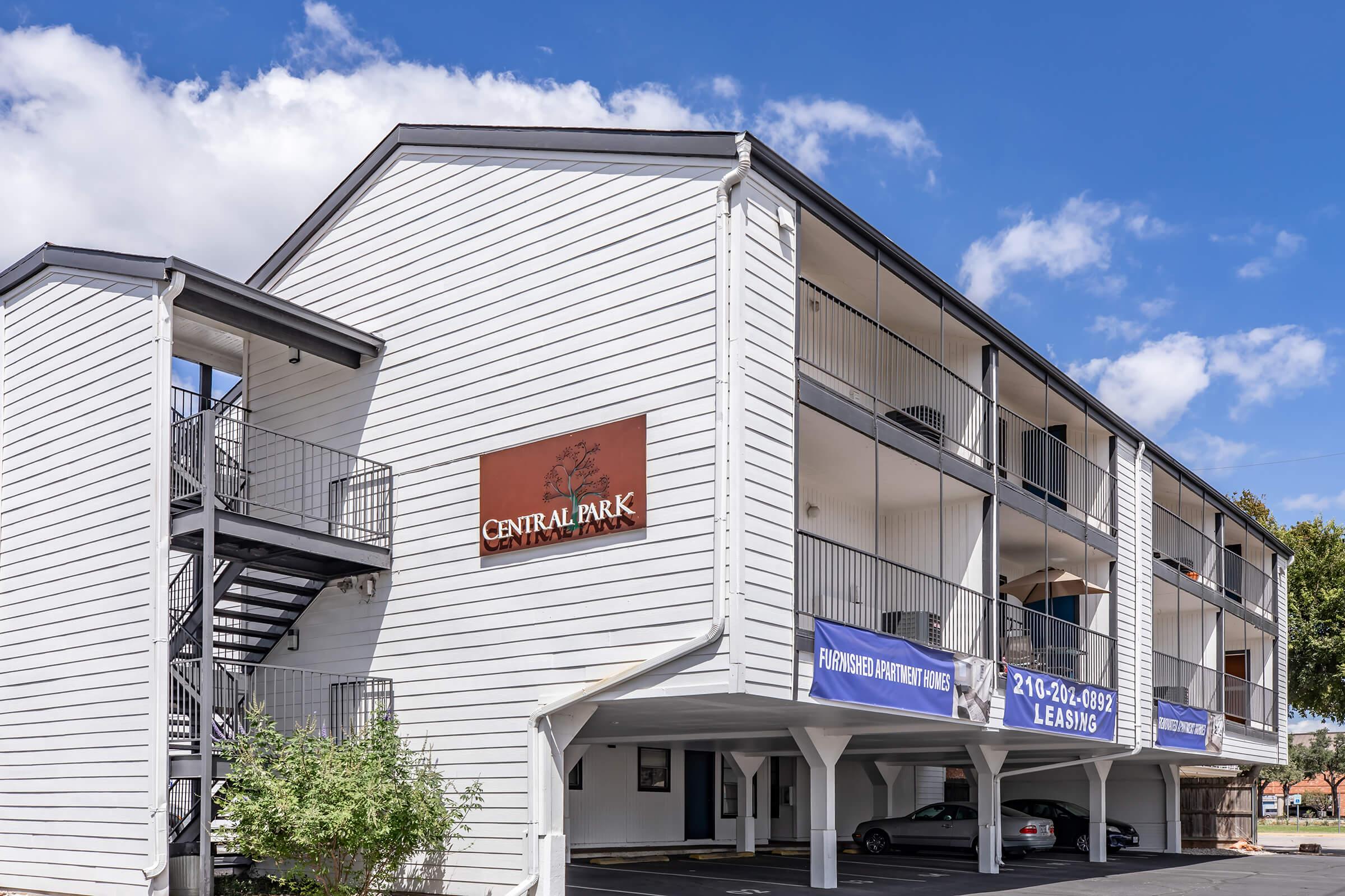 A two-story apartment building with a white exterior featuring multiple balconies. The building has a sign that reads "Central Park" and advertising banners for leasing and contact information. The sky is blue with a few clouds, and there are trees nearby.