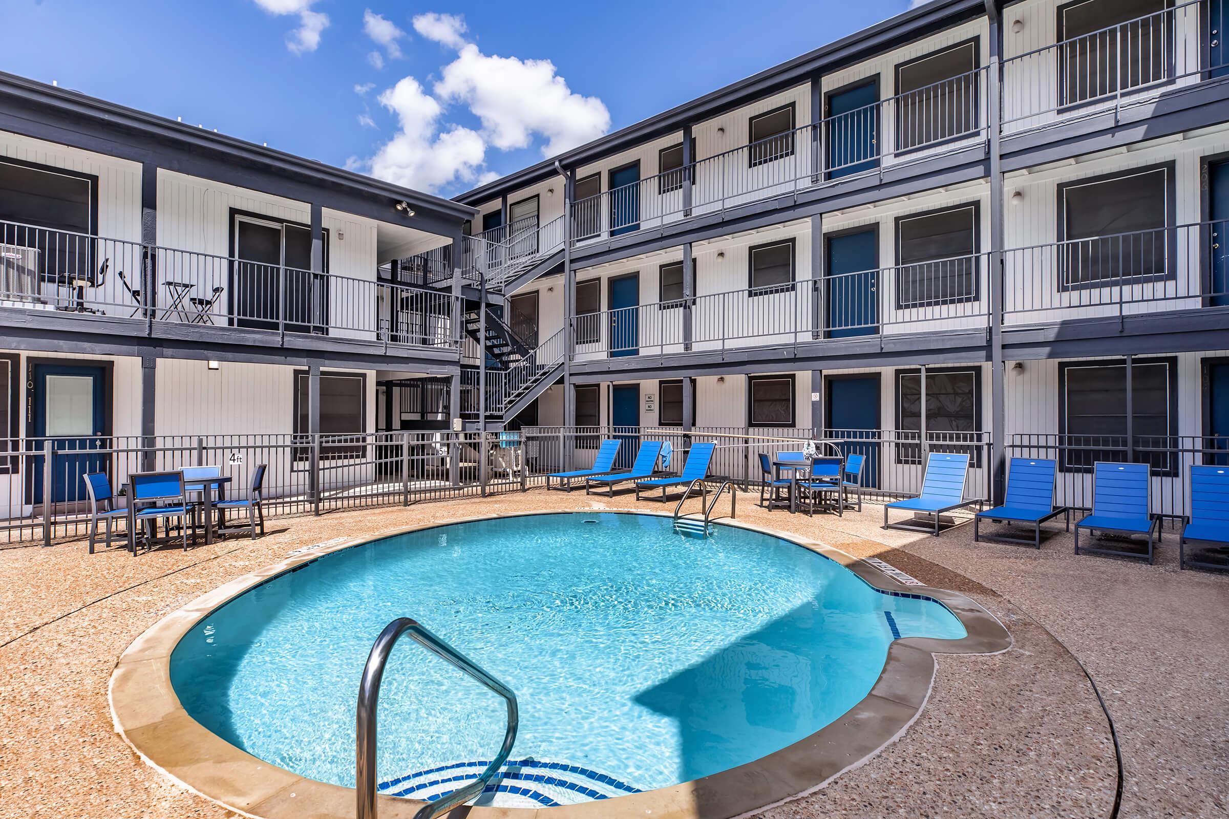 A clear blue swimming pool surrounded by lounge chairs and a patio area. The pool is located in the center of a two-story building with multiple rooms, featuring a modern design and a staircase leading to the upper level. Bright blue skies and fluffy clouds are visible above.