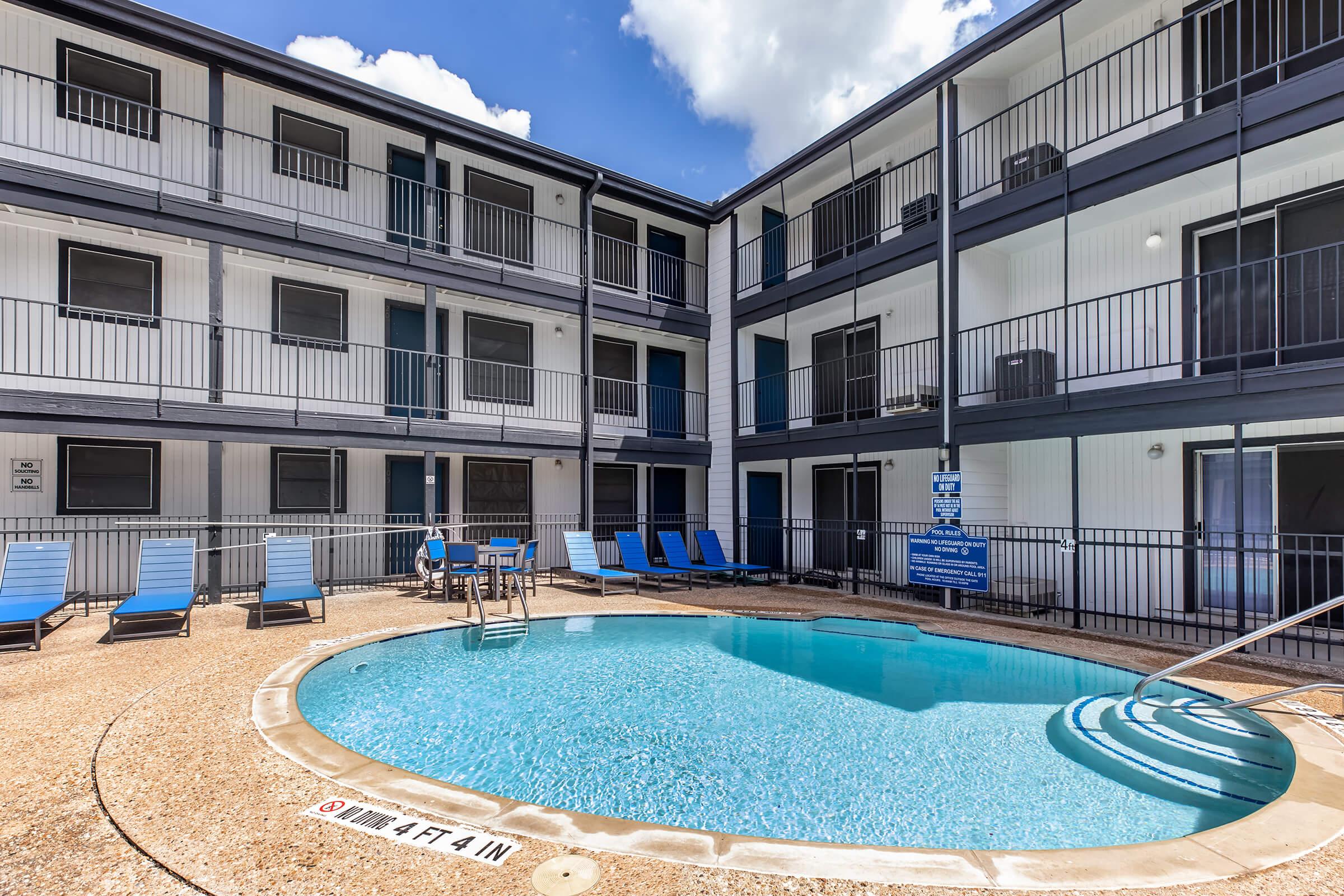 A well-maintained outdoor swimming pool surrounded by lounge chairs, located in a modern, multi-story building with balconies. The pool area features clear water, and there are signs indicating depth. The sky is bright with a few clouds.