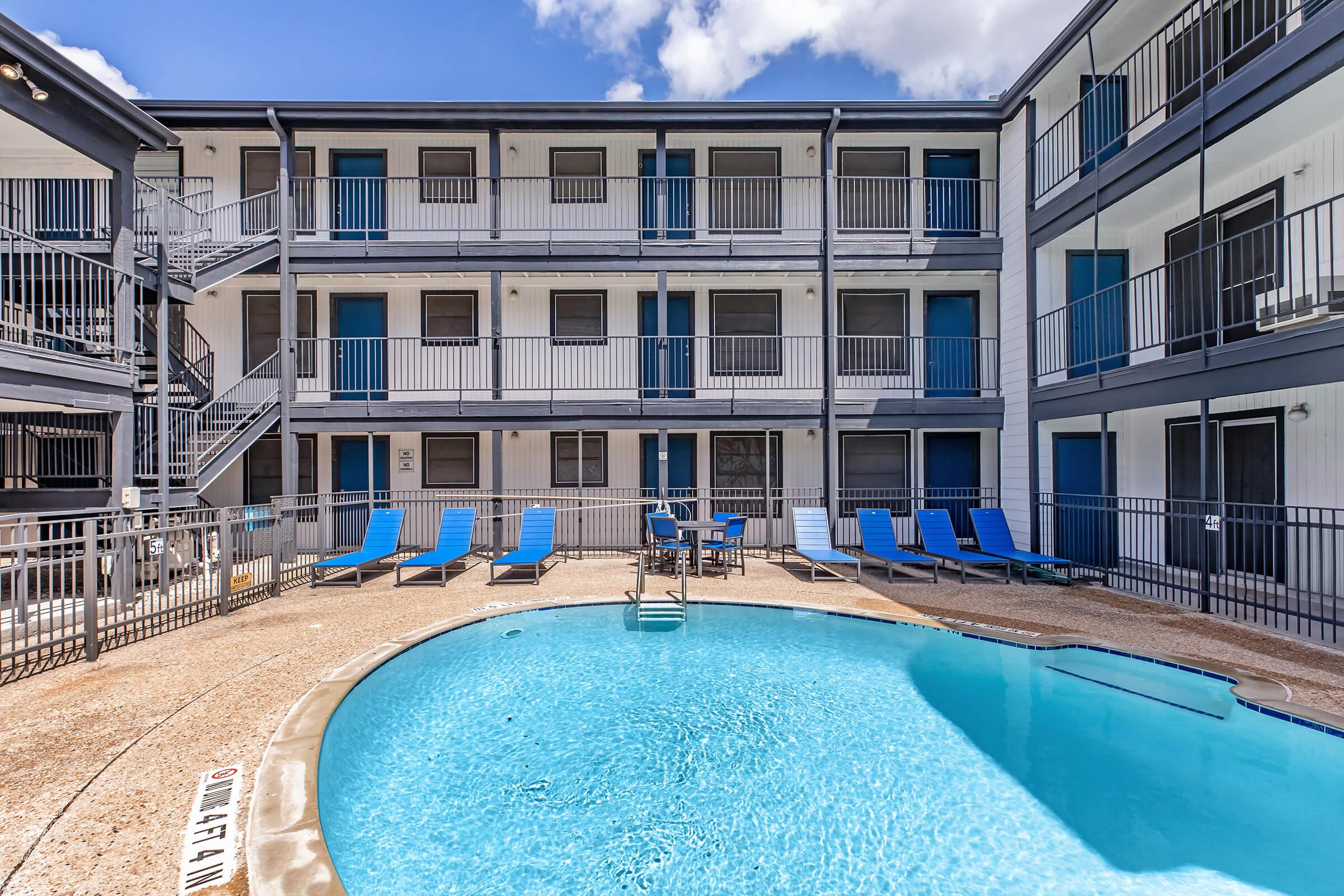 A view of a swimming pool surrounded by lounge chairs and an apartment complex. The pool is clear blue, and the building features three levels with balconies. A spiral staircase leads to the upper floors, while the sky is partly cloudy.